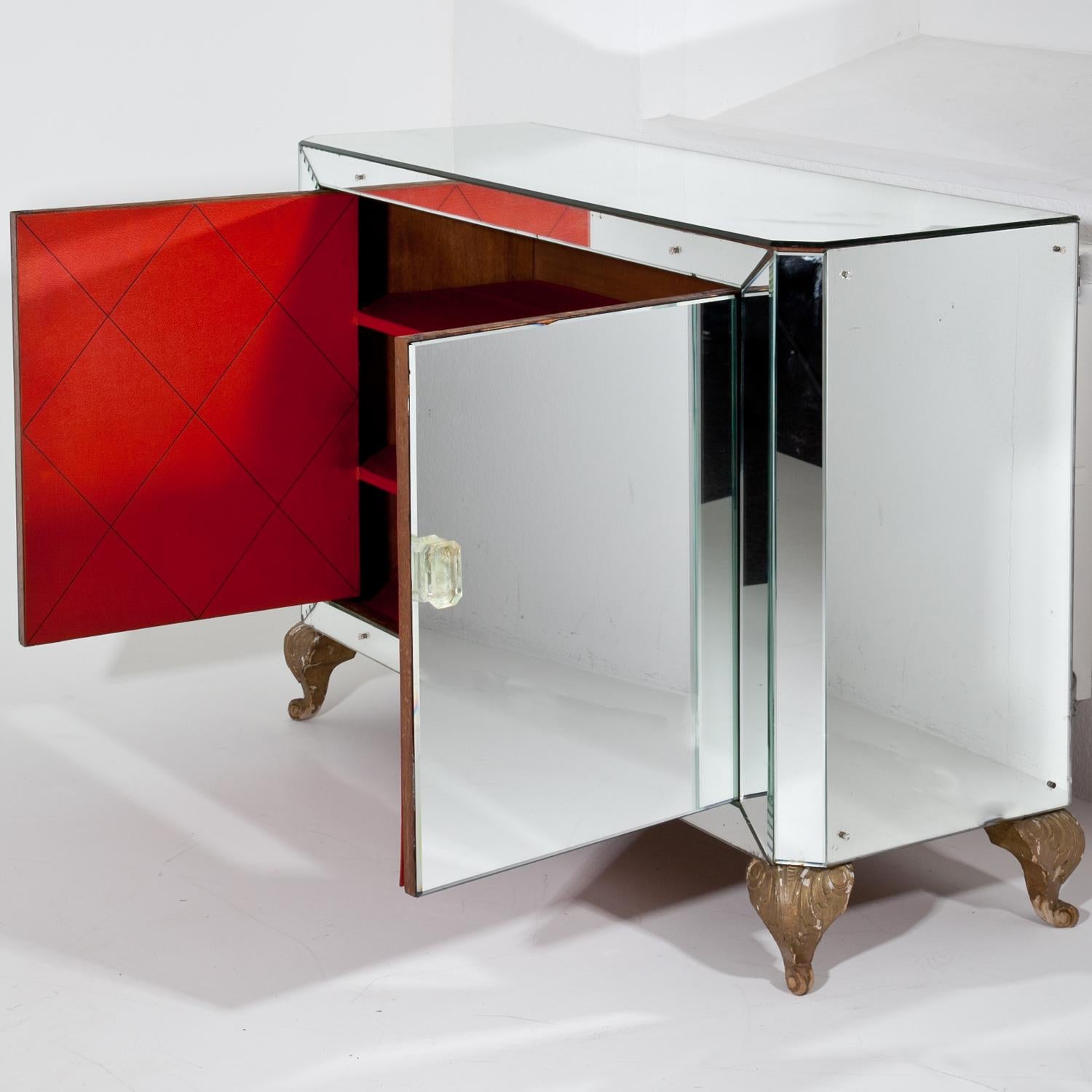 Two-doored bar cabinet, standing on carved volute legs and a completely mirrored body with slanted corners and flower-shaped screw heads. The handles are glass and the insides of the cabinet doors are covered with a red fabric with a rhomboid