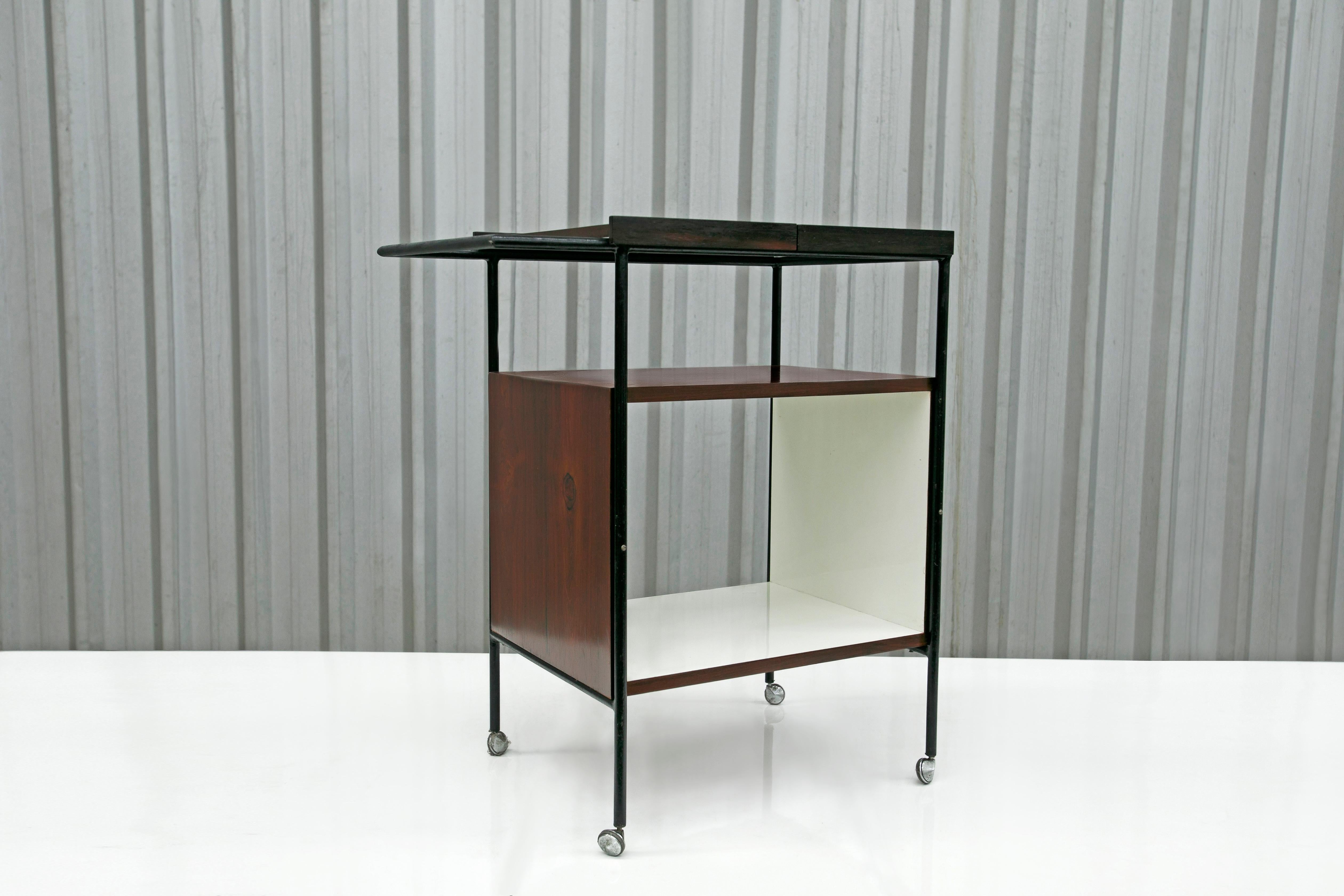 This mid-century modern bar cart is made with Brazilian hardwood and has a black iron frame with white formica on the interior shelf. It features a large shelf, silver wheels on each leg, and two removable trays that can be moved on a sliding