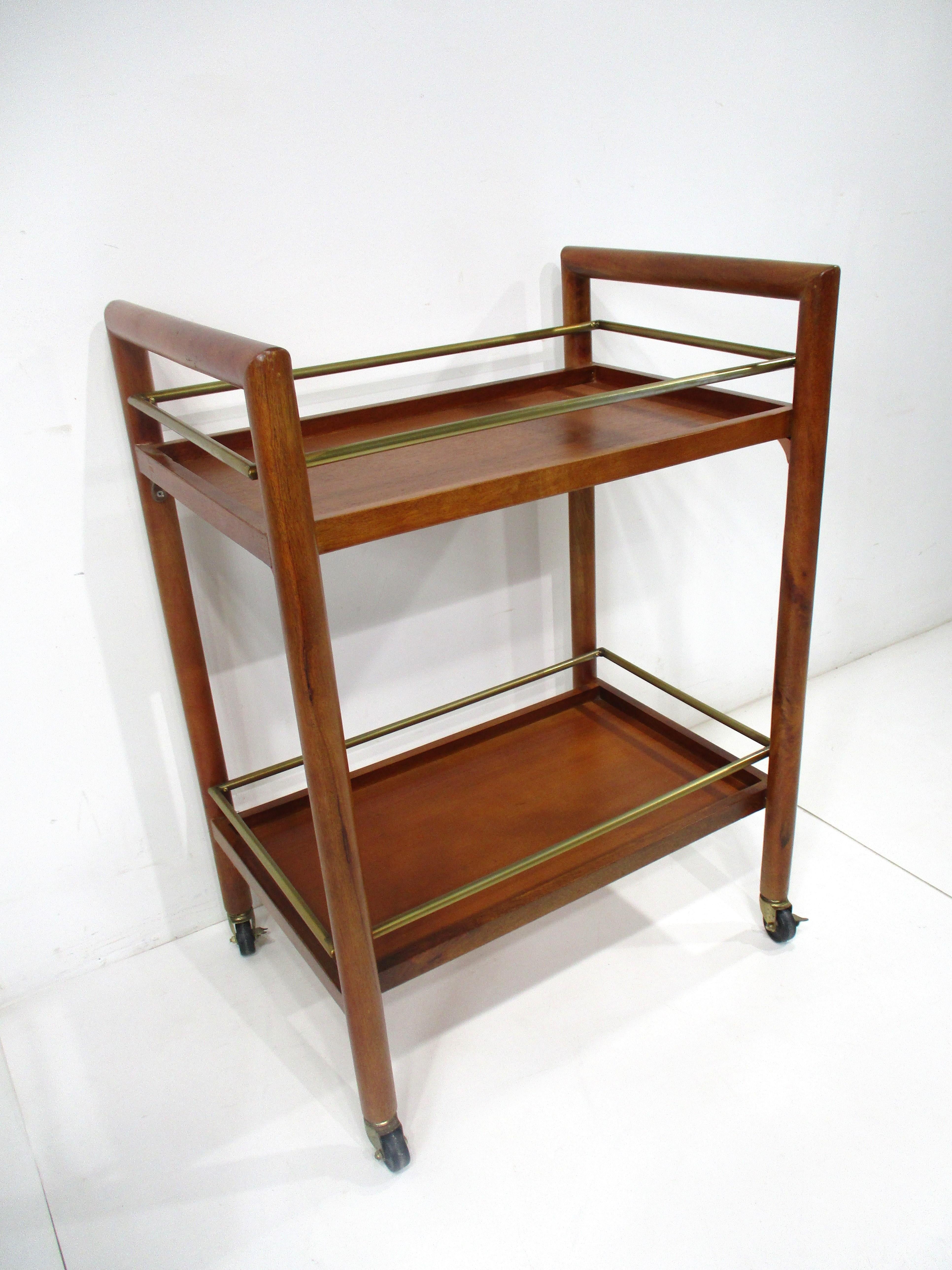 A Mid century mahogany framed rolling bar cart manufactured in the manner of the Widdicomb furniture company and T.H. Robs John - Gibbings . This two tiered cart has polished brass rails to the edge of each shelve making for a nice sophisticated