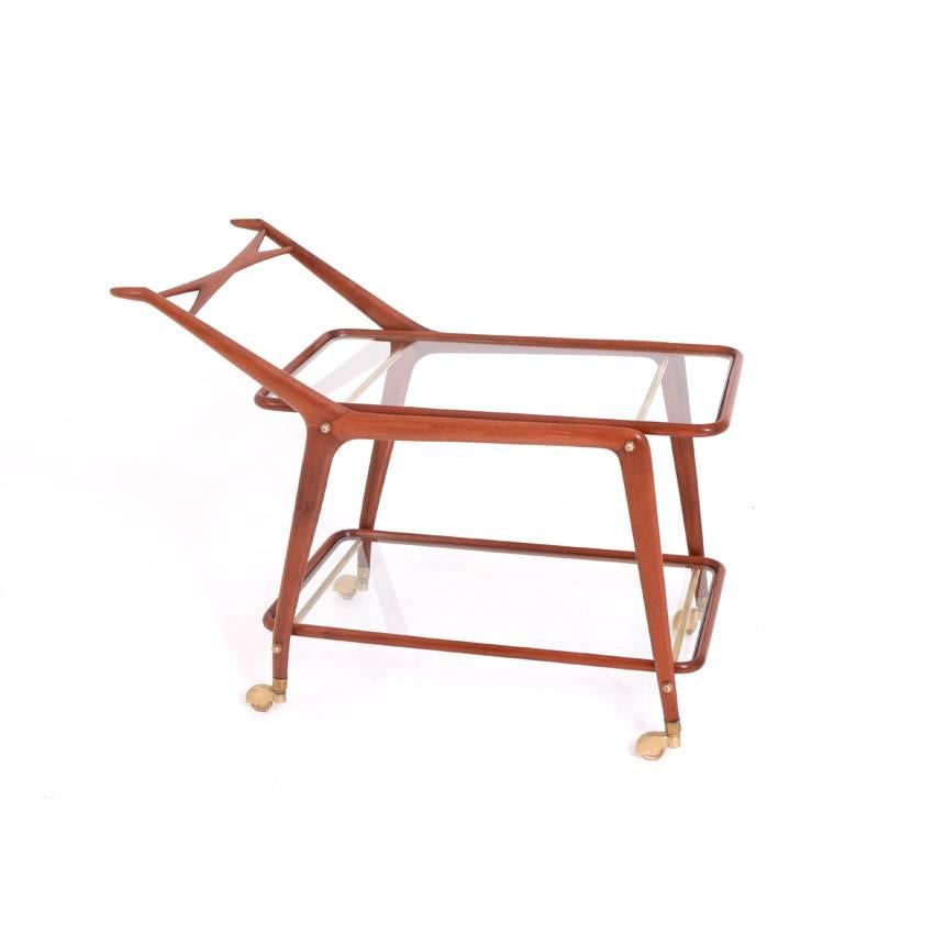 Bar cart on brass castors with two class shelves and a smooth wooden frame with cross-shaped and tapered handles in the style of Ico Parisi or Cesare Lacca.