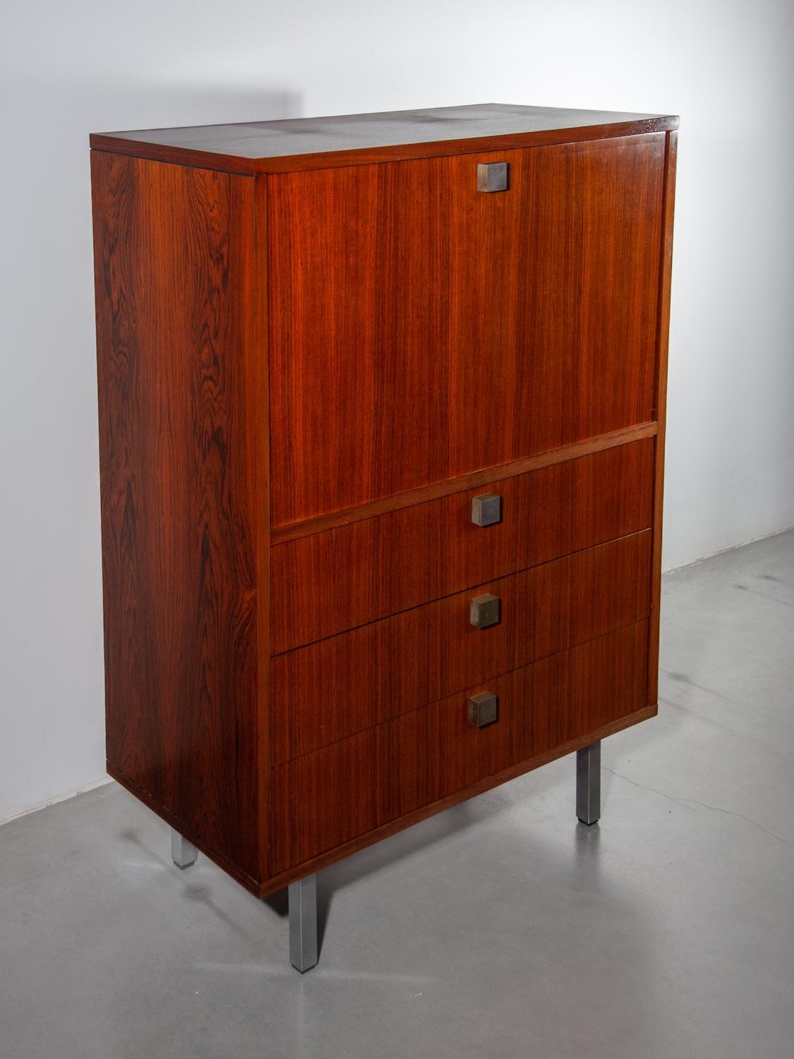 A Bar cabinet, 1960 from the Architect Alfred Hendrickx. A cubic cabinet raised on  silver metal legs, cleverly stores a concealed secretary desk. An upper door folds down to reveal a writing desk, lower three drawers open. The wood veneer is bright
