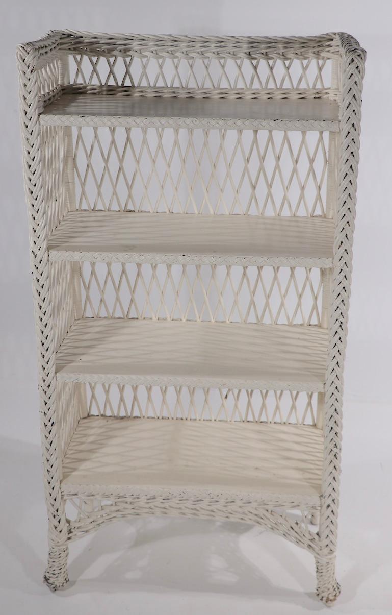 Charming bar Harbor style wicker bookshelf, in white paint 
 This example has four shelves with cross hatched wicker sides and back, braid woven wicker trim and Pineapple front feet. Nice usable scale, good solid condition, clean and ready to use.