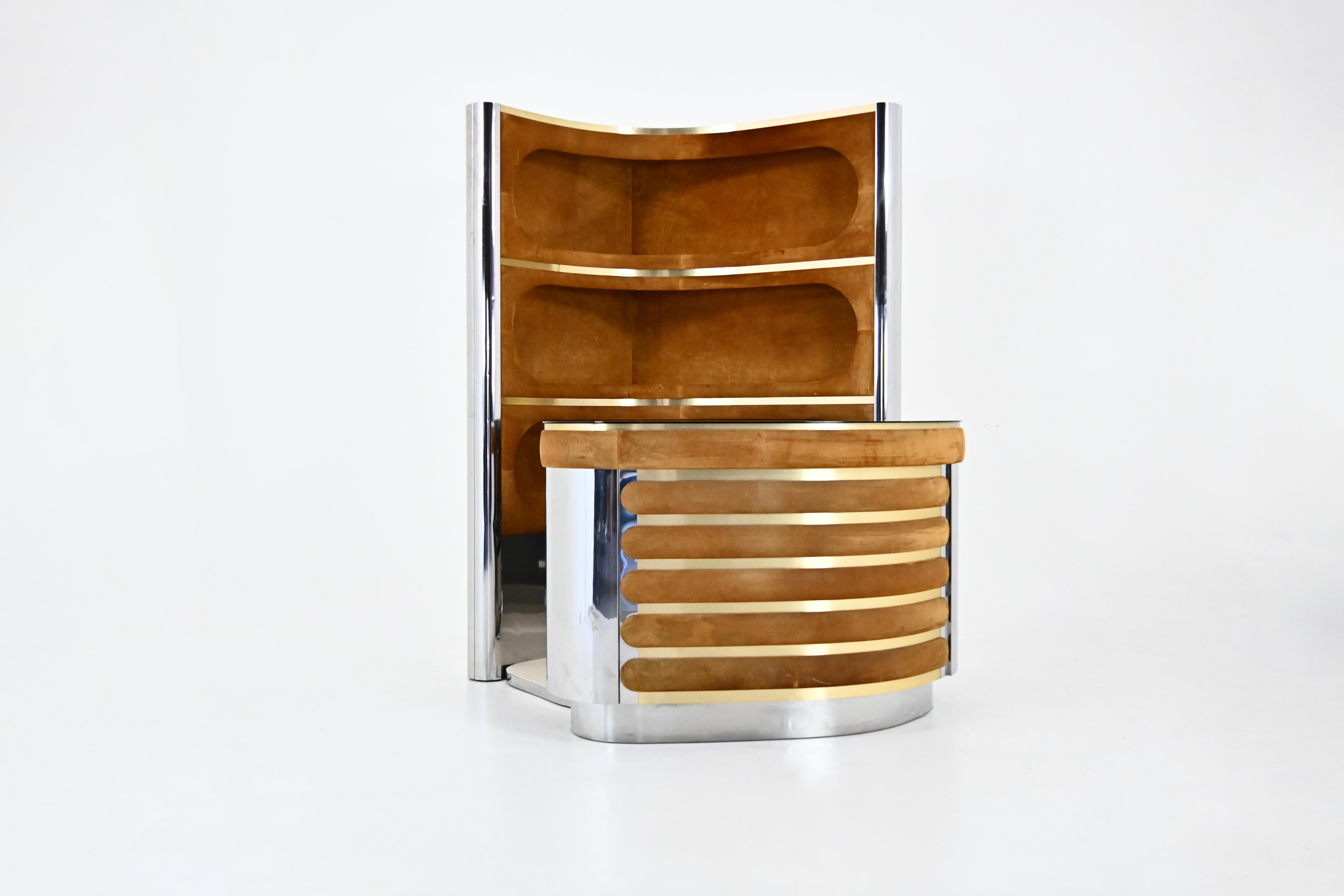 The set includes a cabinet with 3 illuminated shelves in suede, chromed metal and brass.  It also includes a counter in suede, chromed metal with a mirrored top, a fridge and a glass cabinet with a shelf . There is a carpeted floor. 
Shelf