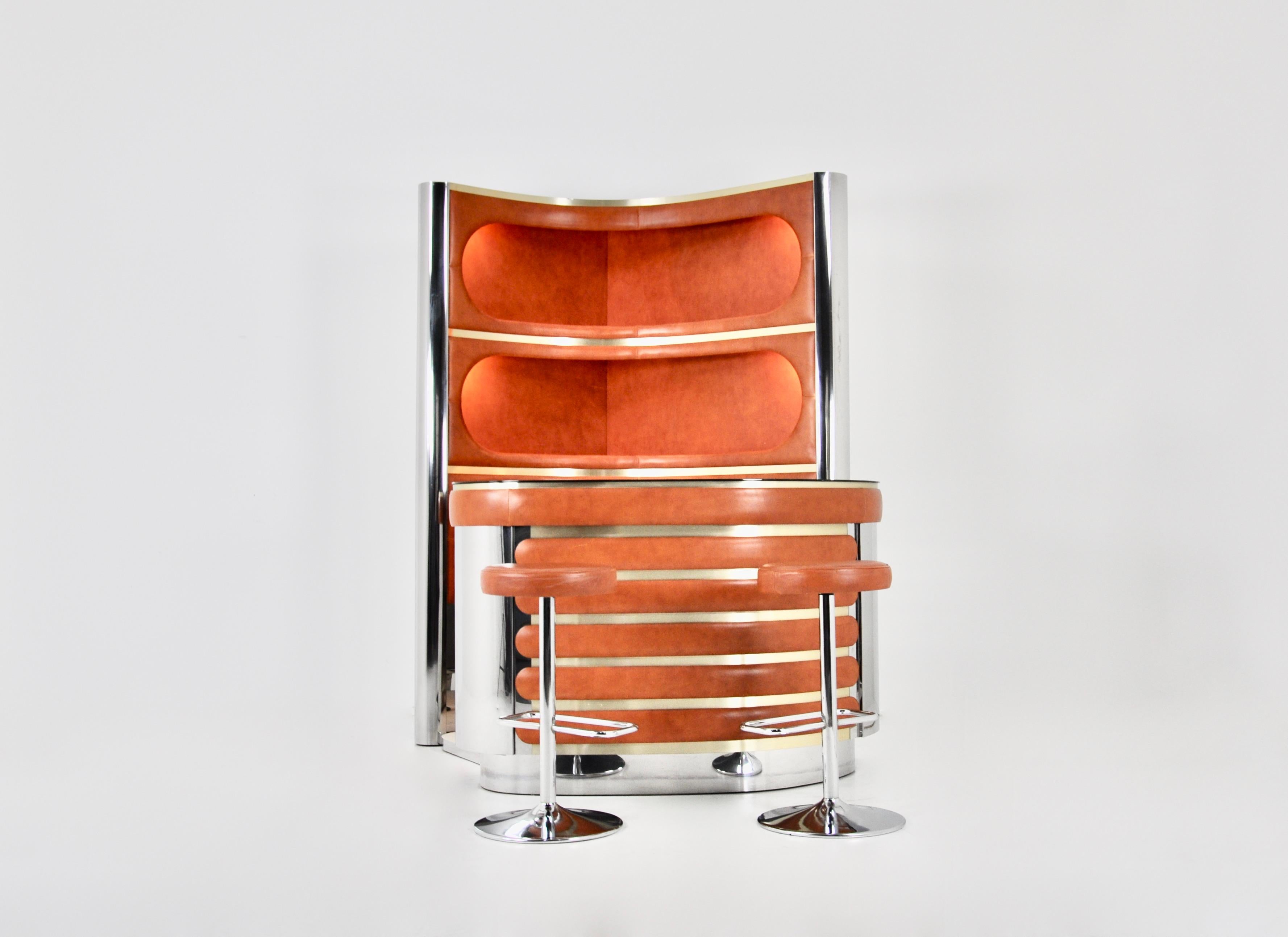 The set includes a cabinet with 3 illuminated shelves in leather, chromed metal and brass.  It also includes a counter in leather, chromed metal, brass with a mirrored top, a fridge and a glass cabinet with a shelf. There is a carpeted floor. 2