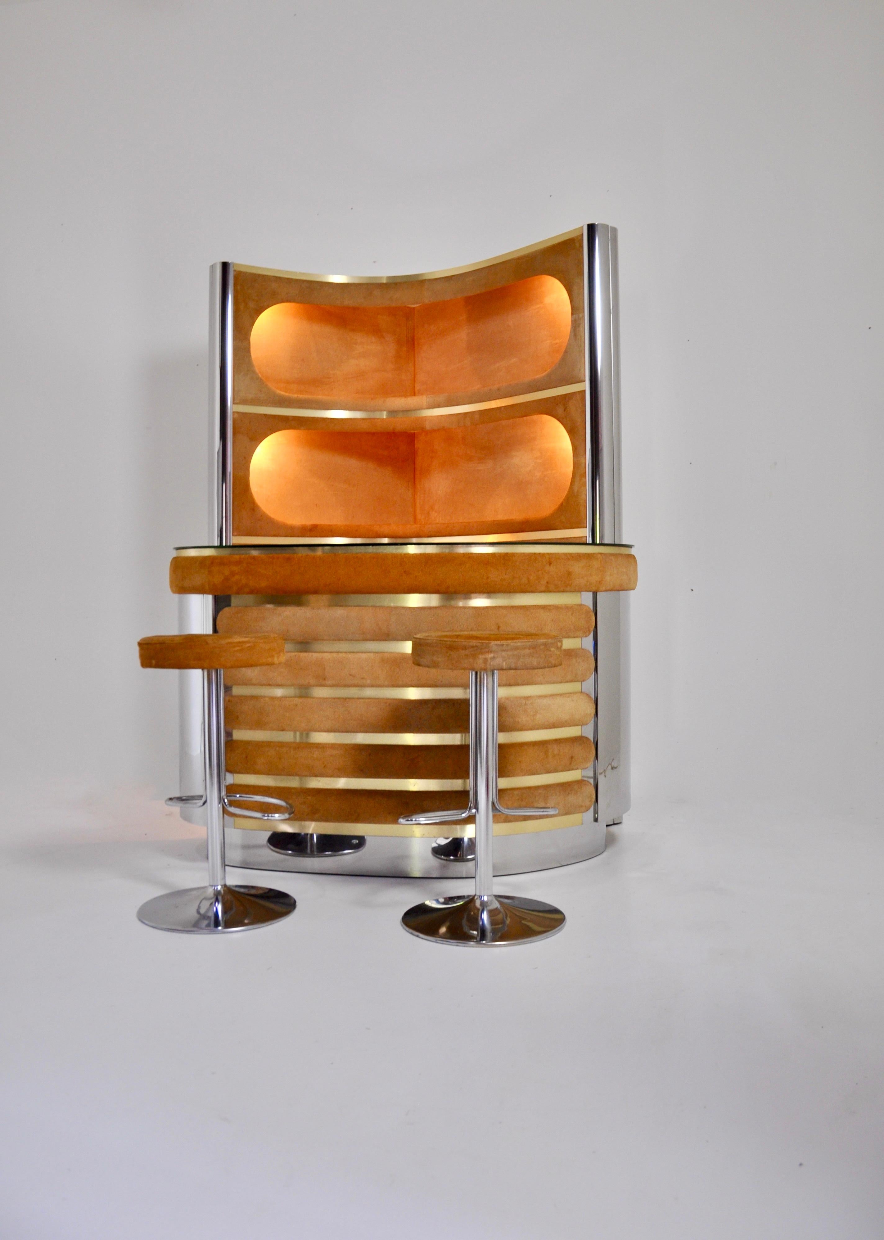 The set includes a cabinet with 3 illuminated shelves in suede, chromed metal and brass.  It also includes a counter in suede, chromed metal with a mirrored top, a fridge and a glass cabinet with a shelf. There is a carpeted floor. 
Stools