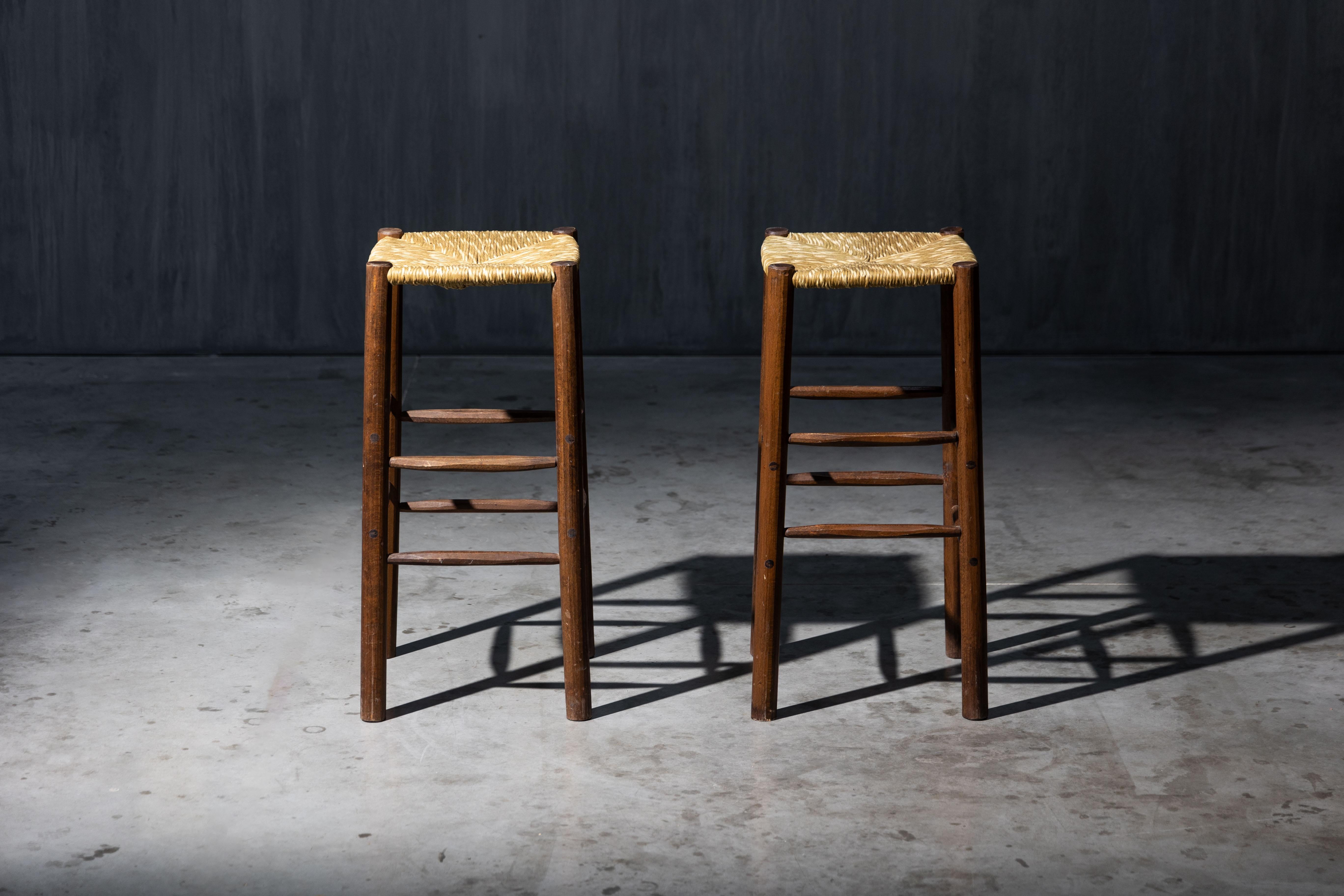 1970s bar stools with woven rush seats, crafted in the timeless style of Charlotte Perriand. Their minimalist wooden frames and natural materials evoke the rustic elegance of French modernism. Both stools are in excellent original condition,