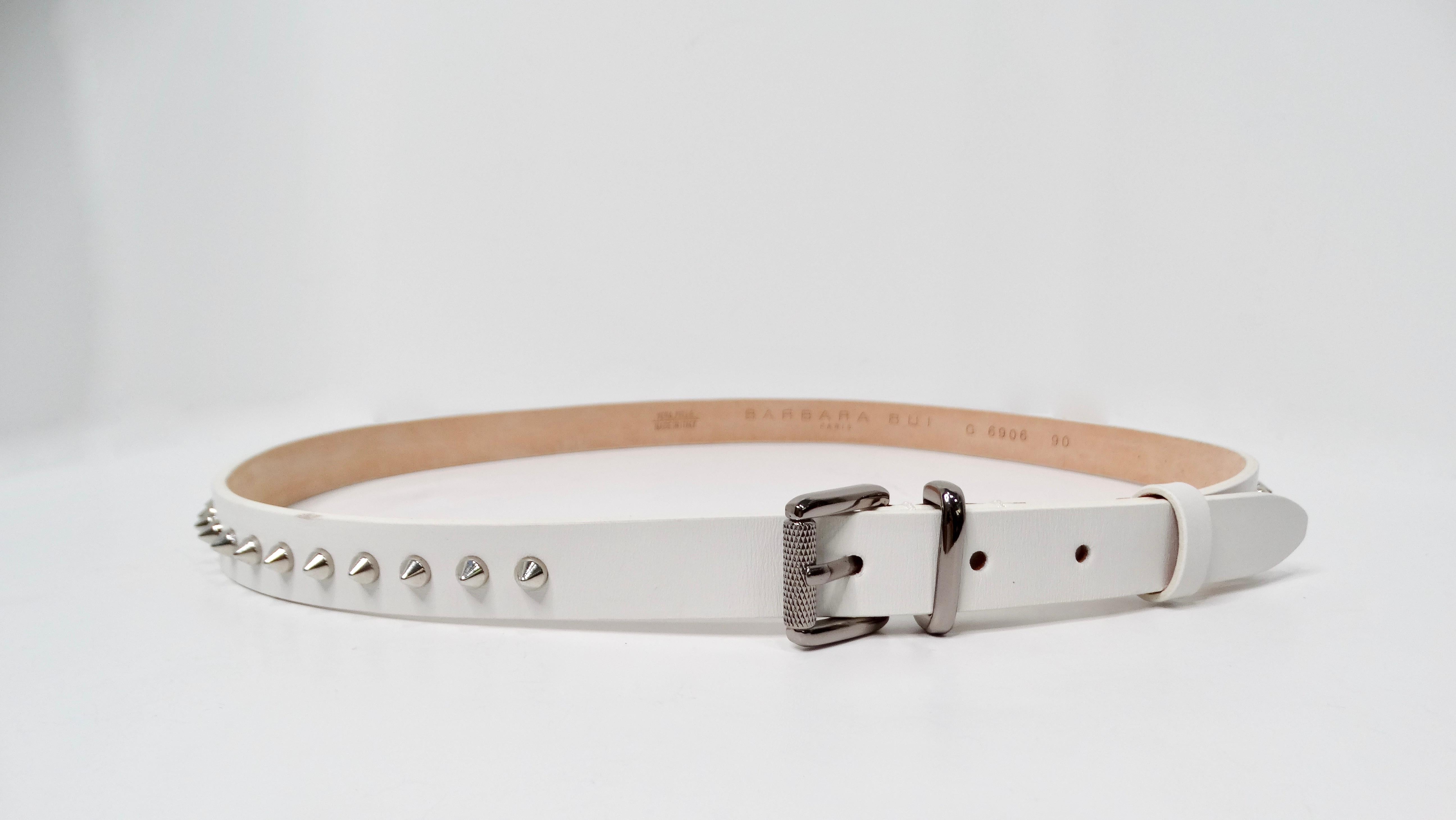 The Barbara Bui White Leather Silver Stone Studded Belt is a quintessential accessory that seamlessly blends elegance with an edgy twist. This skinny belt features a white leather base adorned with striking silver-tone studs, adding a touch of