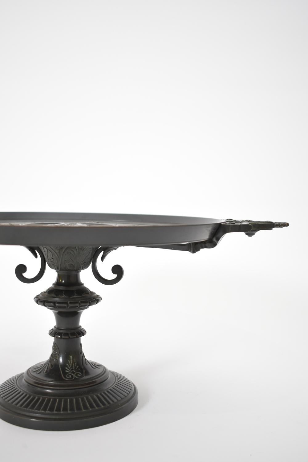 Plat de service, coupe sur piédouche, en bronze, patine ancienne, le plateau orné en son centre d'un profil d'Athéna entouré d'une frise de coquillages, guirlandes, rubans, rosaces et volutes, prise latérale à décor de palmettes. La tige est ciselée
