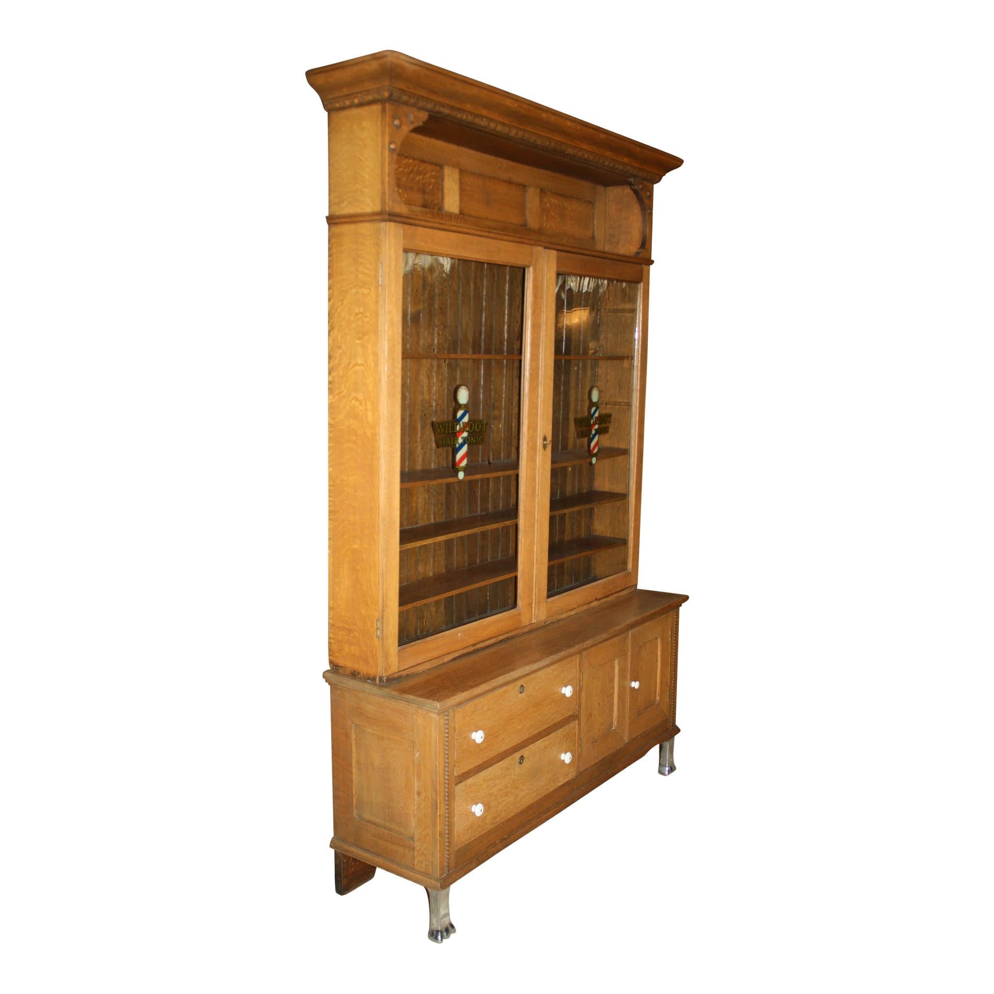 This two piece, vintage, oak cabinet was used to display wildroot hair products in a barbershop or store in the mid-20th century. The top features a recessed niche above two glass doors, which open to four removable shelves. The bottom is comprised