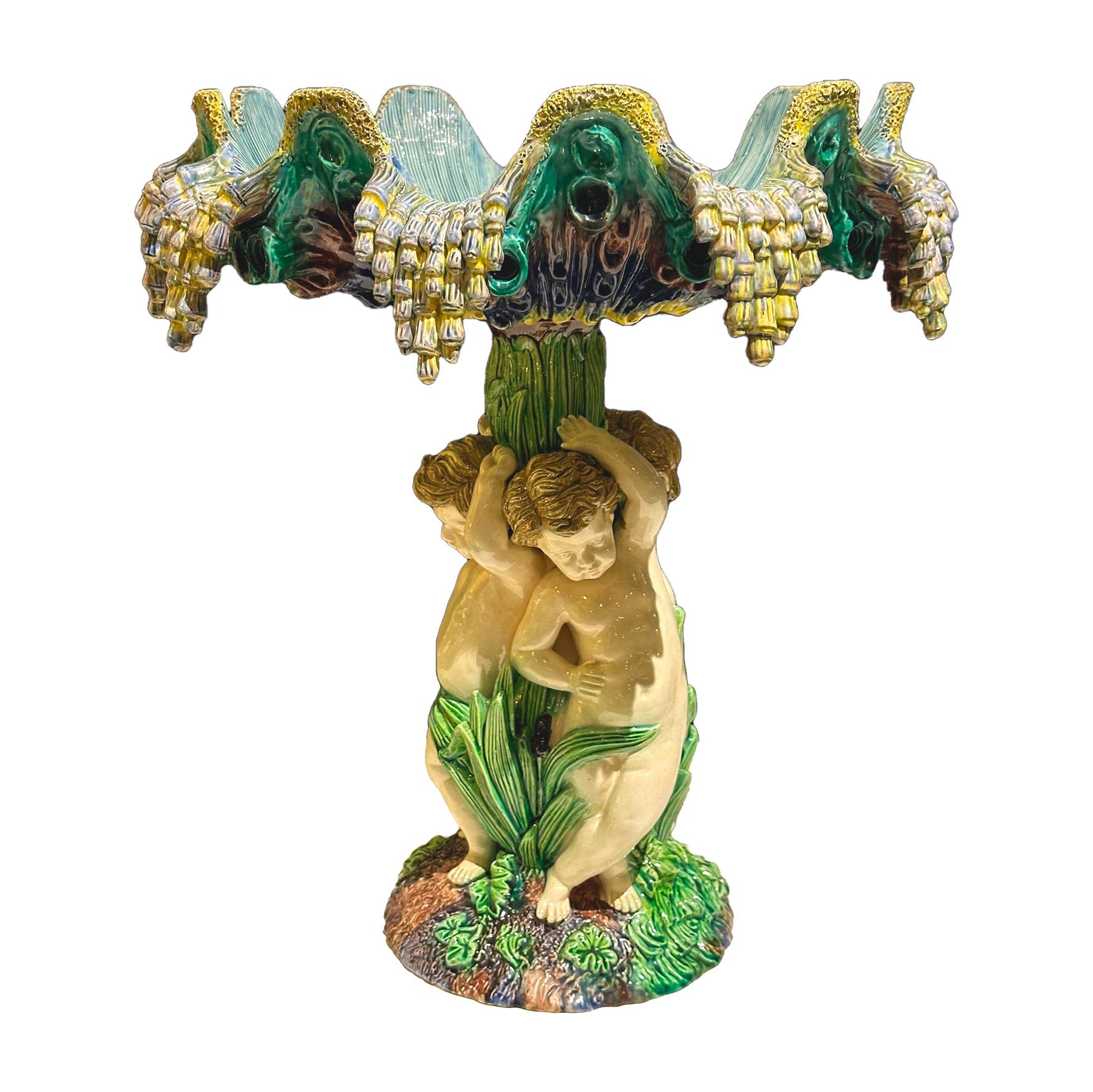 Cet objet est un centre de table en Barbotine, une technique de céramique où une pâte liquide est moulée, émaillée et cuite pour obtenir un aspect coloré et texturé. L'œuvre représente trois chérubins (ou putti), petits personnages nus souvent