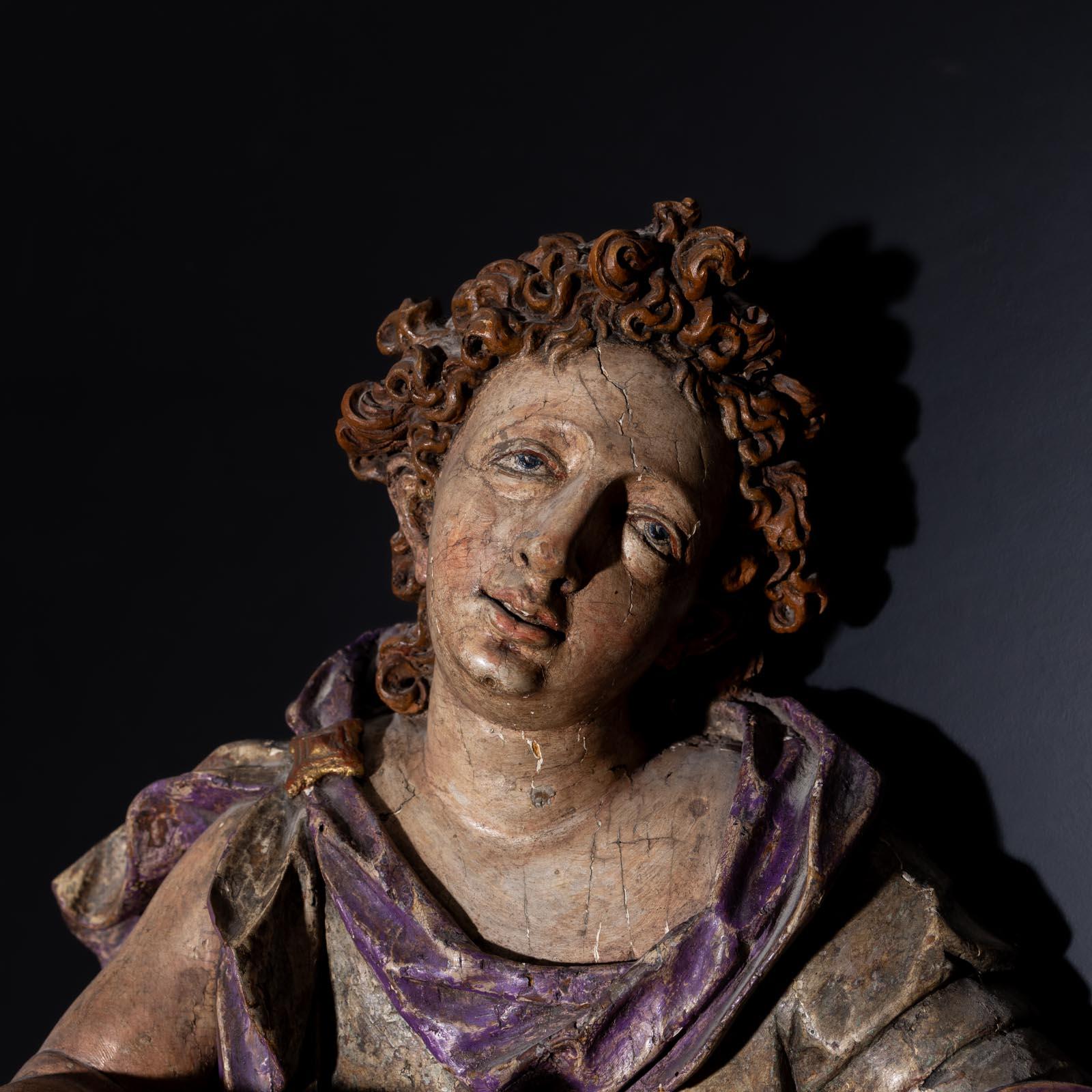 Large baroque wall sculpture in the shape of an angel with curly hair and a transfigured gaze. He holds a scroll in his hands and sits on a cloud. The polychrome paint is rubbed and chipped in places.