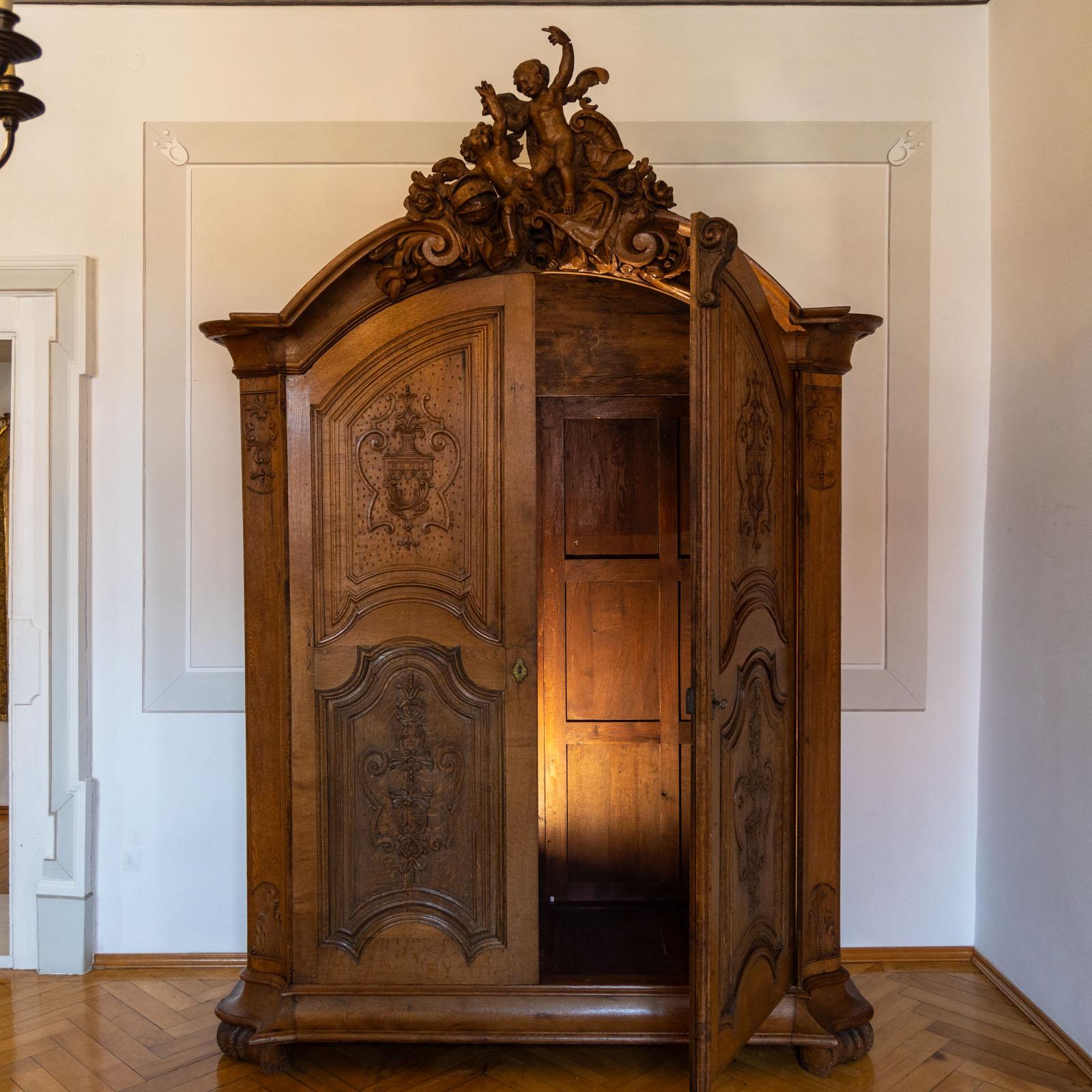 Large baroque hall cupboard made of solid oak with imposing carved and fully figured pediment with winged putti. The cabinet was constructed using old parts. The front is adorned with four panels with a profiled frame and carved amphora decoration.  