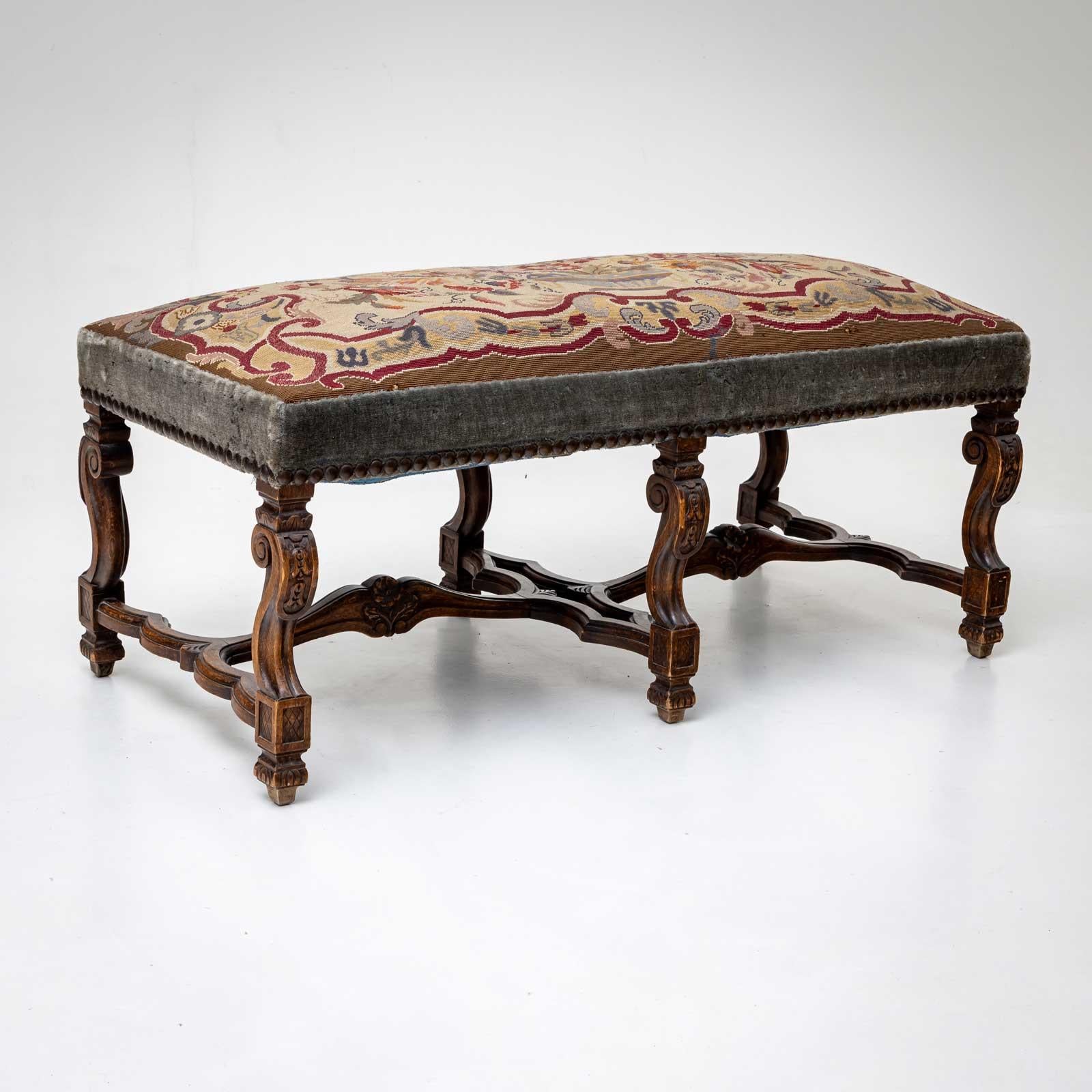 Bench on a carved wooden frame with six scrolled legs and curved braces. The bench is covered with an old tapestry that is riveted all around.