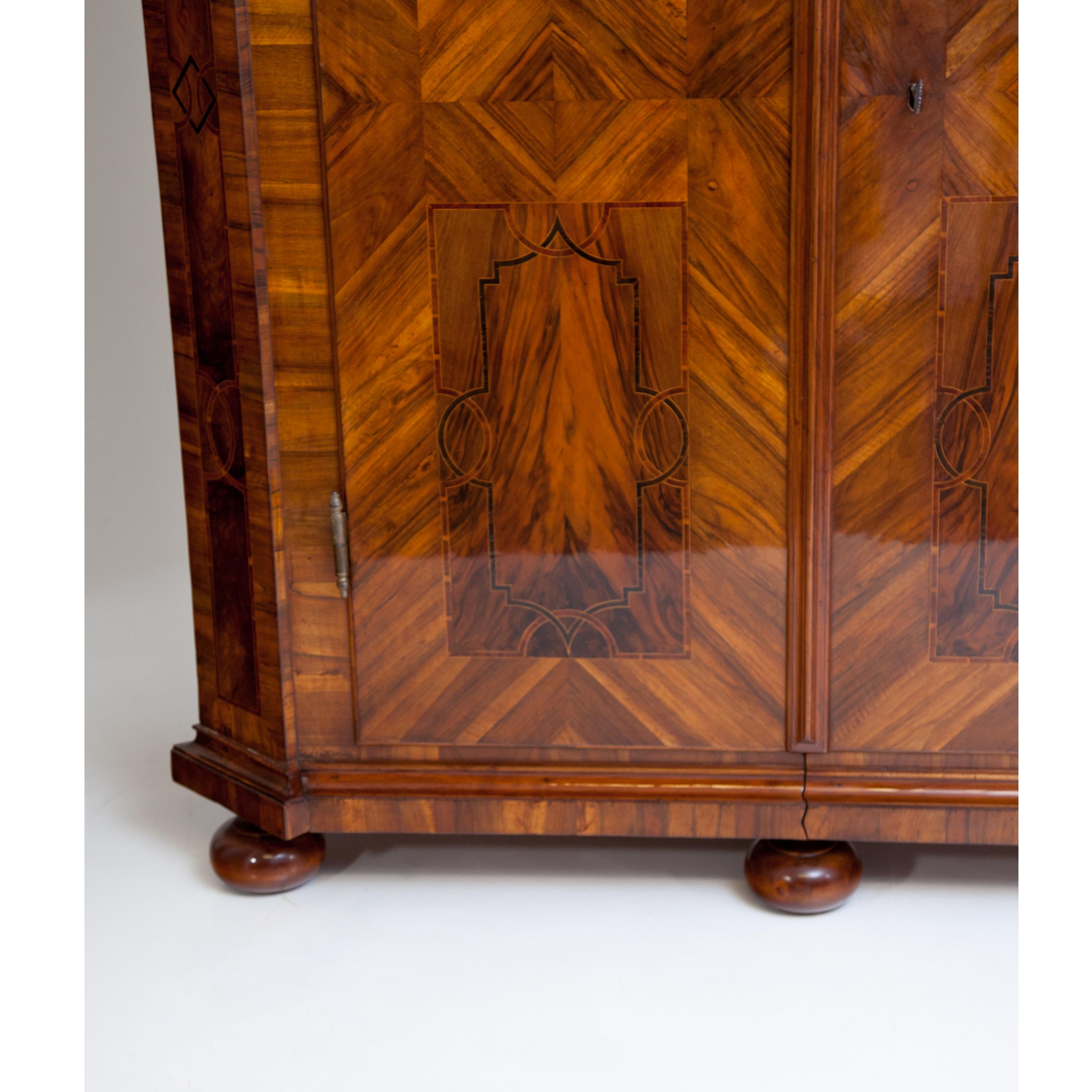 Large two-door Baroque cabinet in walnut veneer, with slightly protruding straight cornice with profiled mouldings and bevelled corners. The cabinet stands on spherical feet and shows ribbon inlays on the corners and the doors divided into four