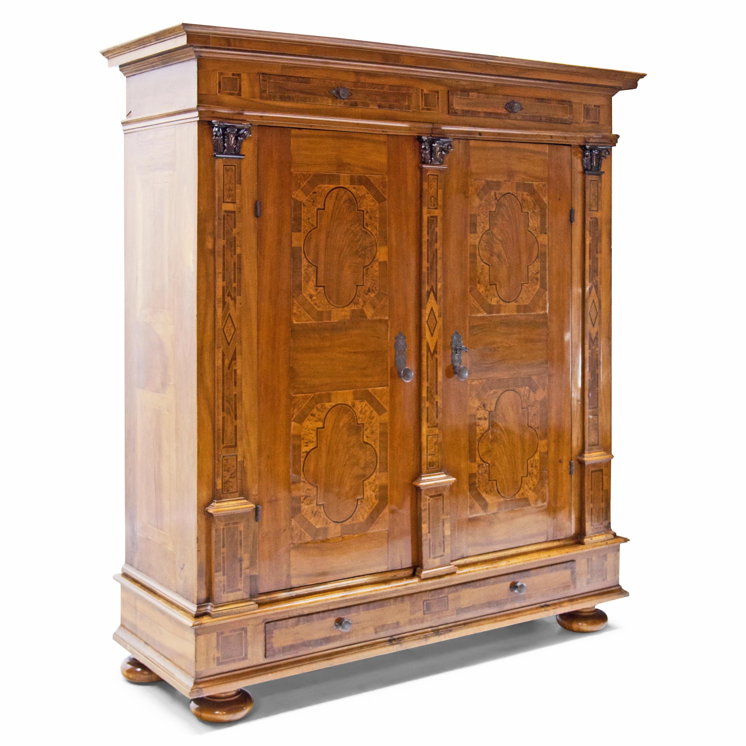 Two-door walnut veneered Baroque cabinet standing on bun feet, the base is fitted with two narrow drawers between profiled strips. Above it the body is divided by three pilasters with carved acanthus leaf capitals and thread or ribbon inlays. The