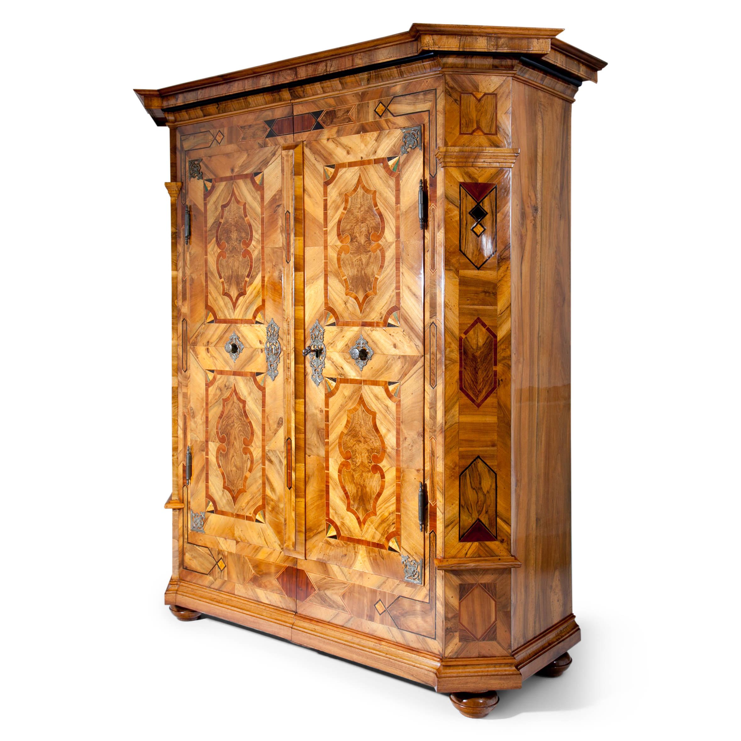 Two-doored baroque cabinet on bun feet with a profiled base and a protruding pediment. The beveled corners are decorated with pilasters. The doors show cast iron fittings in each corner as well as pull handles and escutcheons. Very beautiful walnut