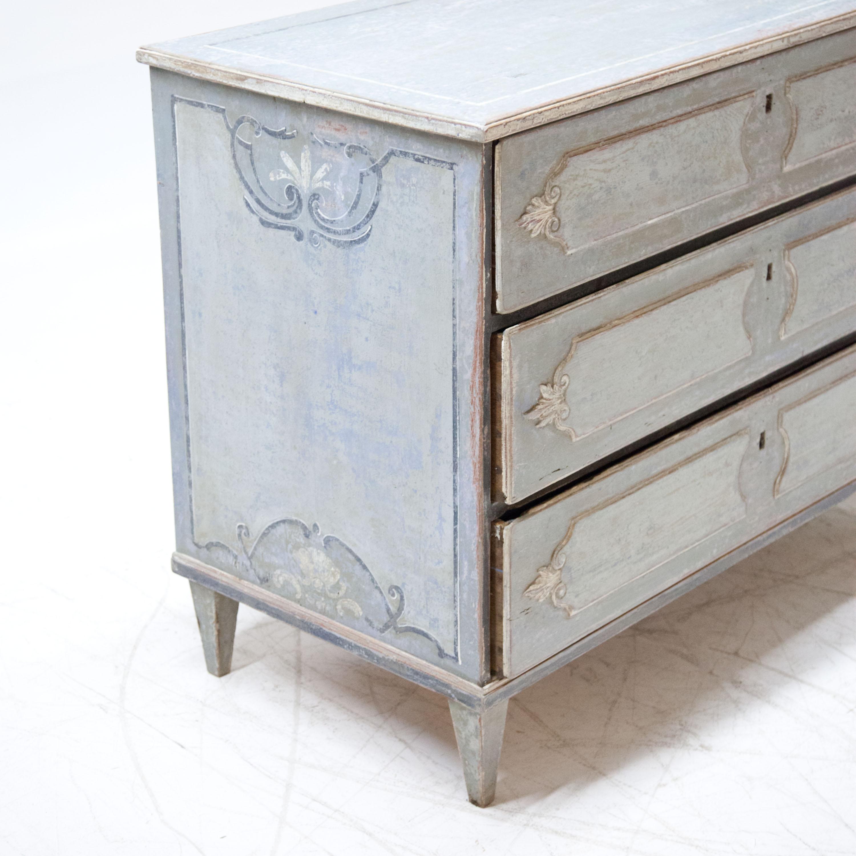 Chest of drawers with three drawers standing on square tapered feet. The body with blue setting with ribbon work and rocaille motifs. The drawers with filling-shaped curved cartouches. The setting is new and made according to historical motifs.