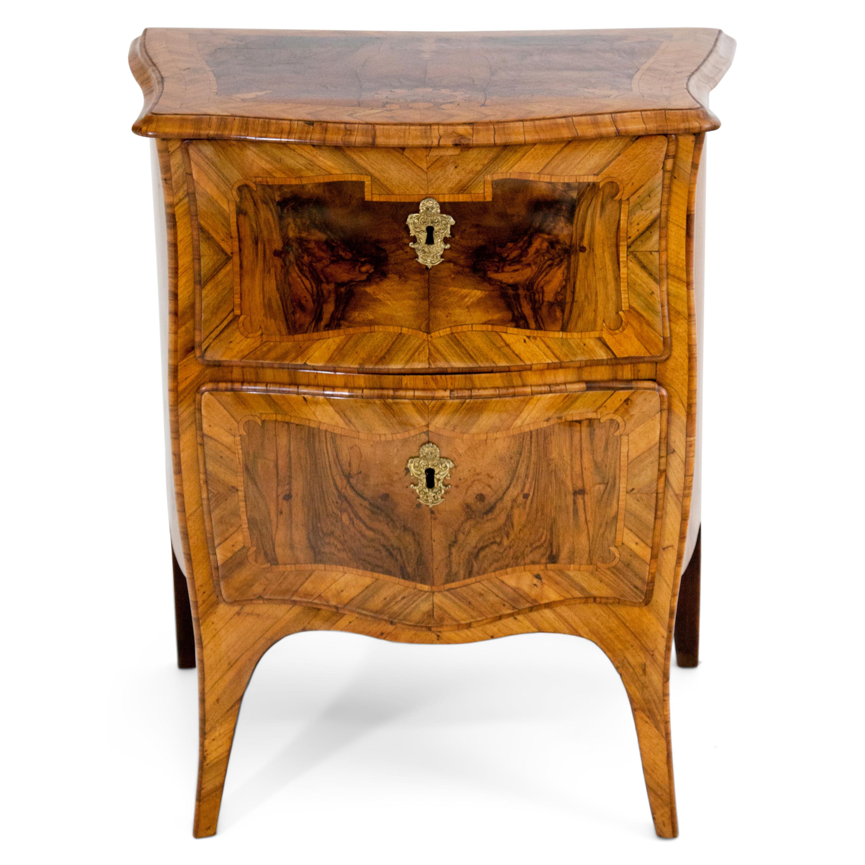Small two-drawered Baroque chest of drawers with a narrow trapezoidal body and bombé front and sides. Walnut veneered with ribbon inlays and rocailles.
  