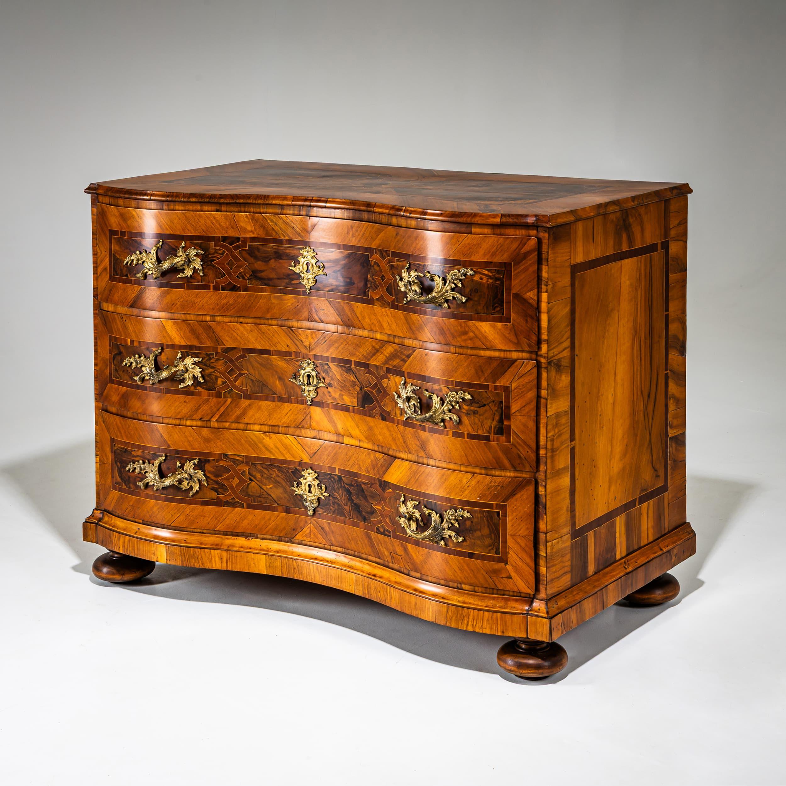 Baroque chest of drawers with three drawers and a serpentine front. Resting on compressed baluster legs, it is fitted with fire-gilt mounts in asymmetrical stylized leaf form. The surfaces feature refined bandwork inlays and walnut panels with dark