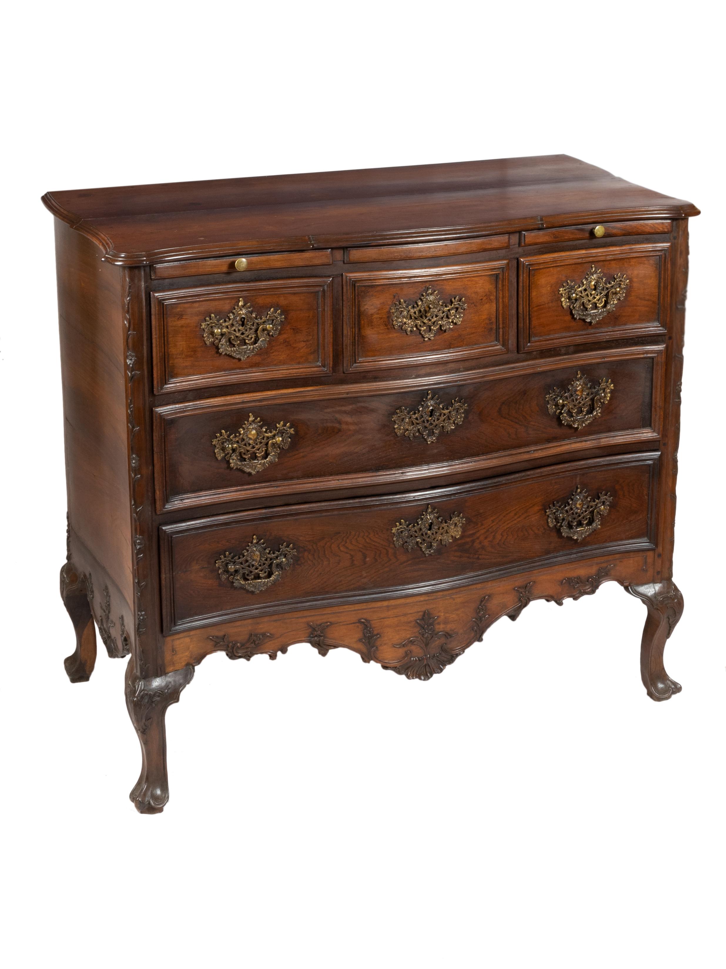 A  richly wooden commode chest of drawers with a scalloped top and corners, two small drawers and three big drawers, cut-out and finely carved skirts with floral motifs and volutes with winding endings. Partially rounded pilasters with well-curved