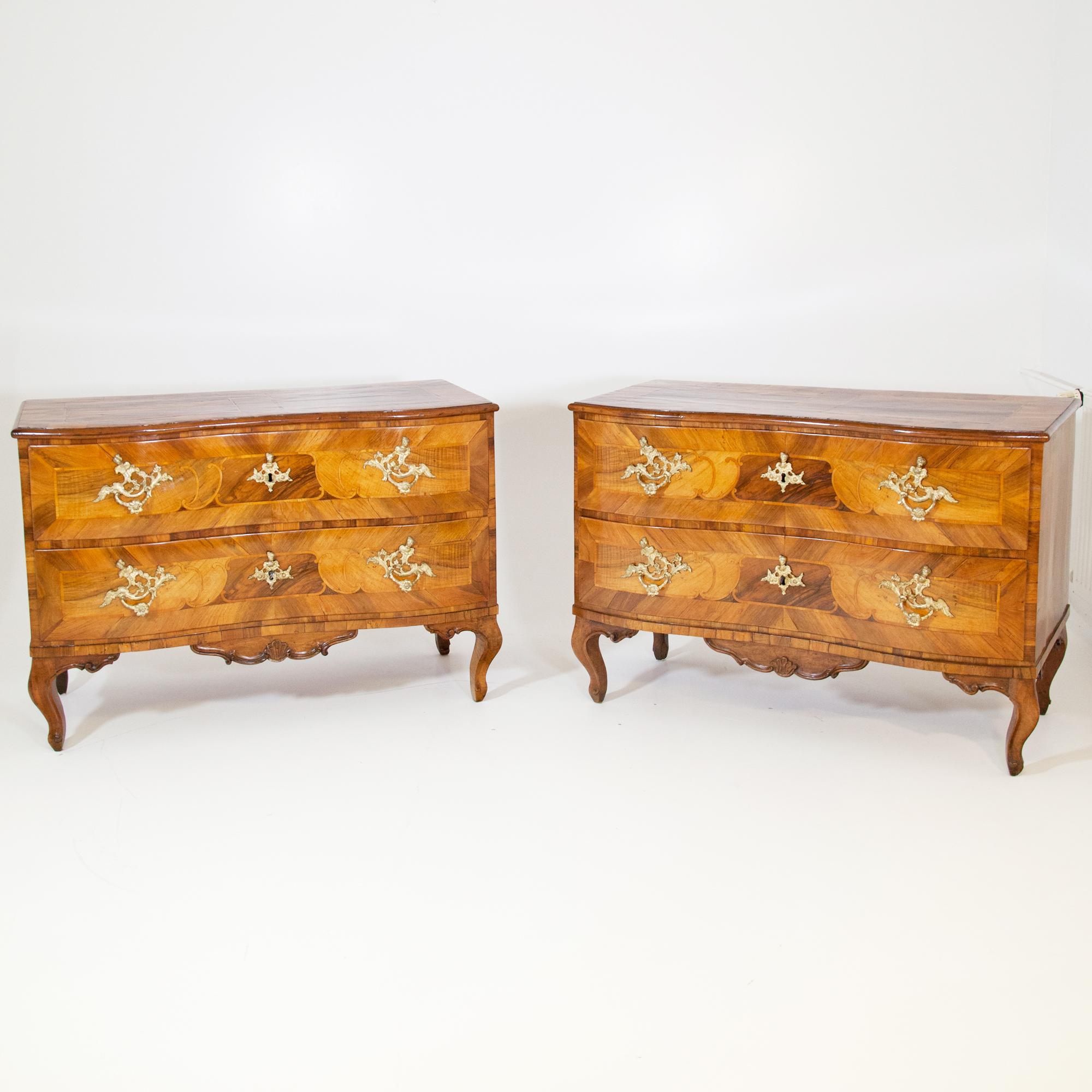 Two-drawered walnut chests of drawers standing on curved legs, with slightly curved fronts and carved aprons with shell motifs. The drawers are decorated with strap work and fire-gilded original bronze fittings. Museum-like, original condition with