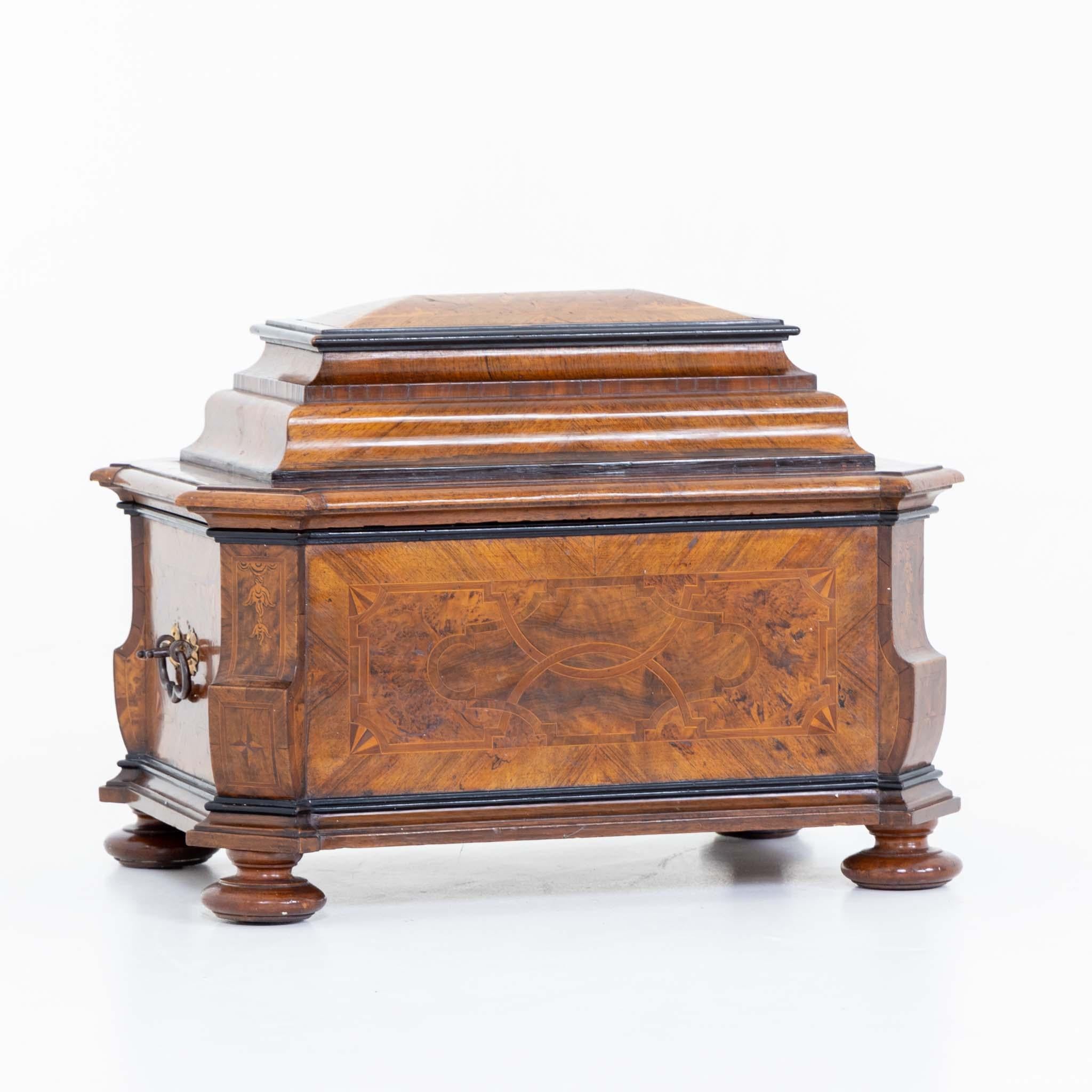 Baroque guild chest on baluster legs in walnut veneer with strap work inlays and side handles. The cushion-shaped lid slides open to reveal the lock.