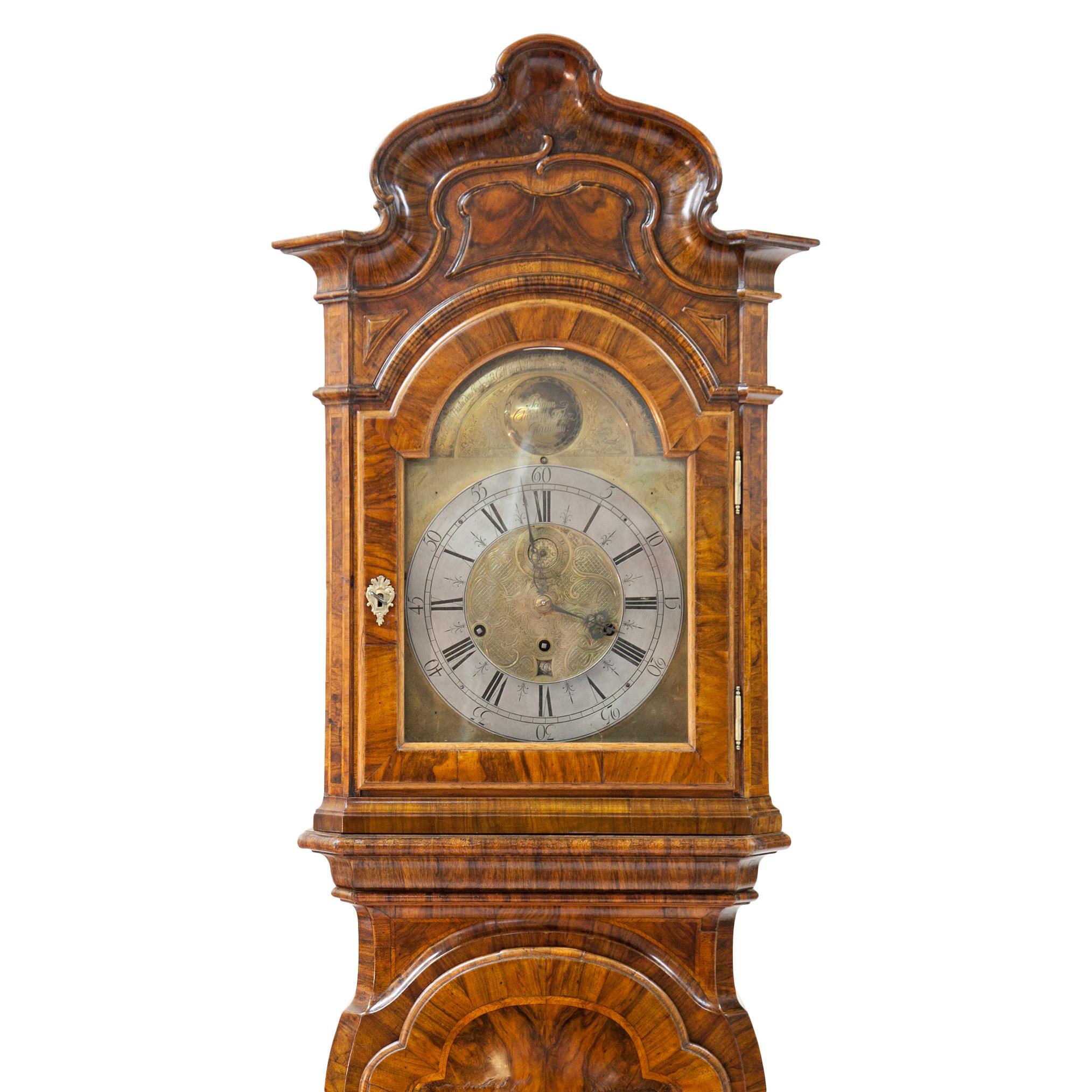 Large baroque grandfather clock in walnut veneered, with a curvy base and a trapezoidal pendulum case. The top part shows a wavy cornice, one glazed door and lateral carved fillings in black. The door with a quatrefoil pendulum window. The clock