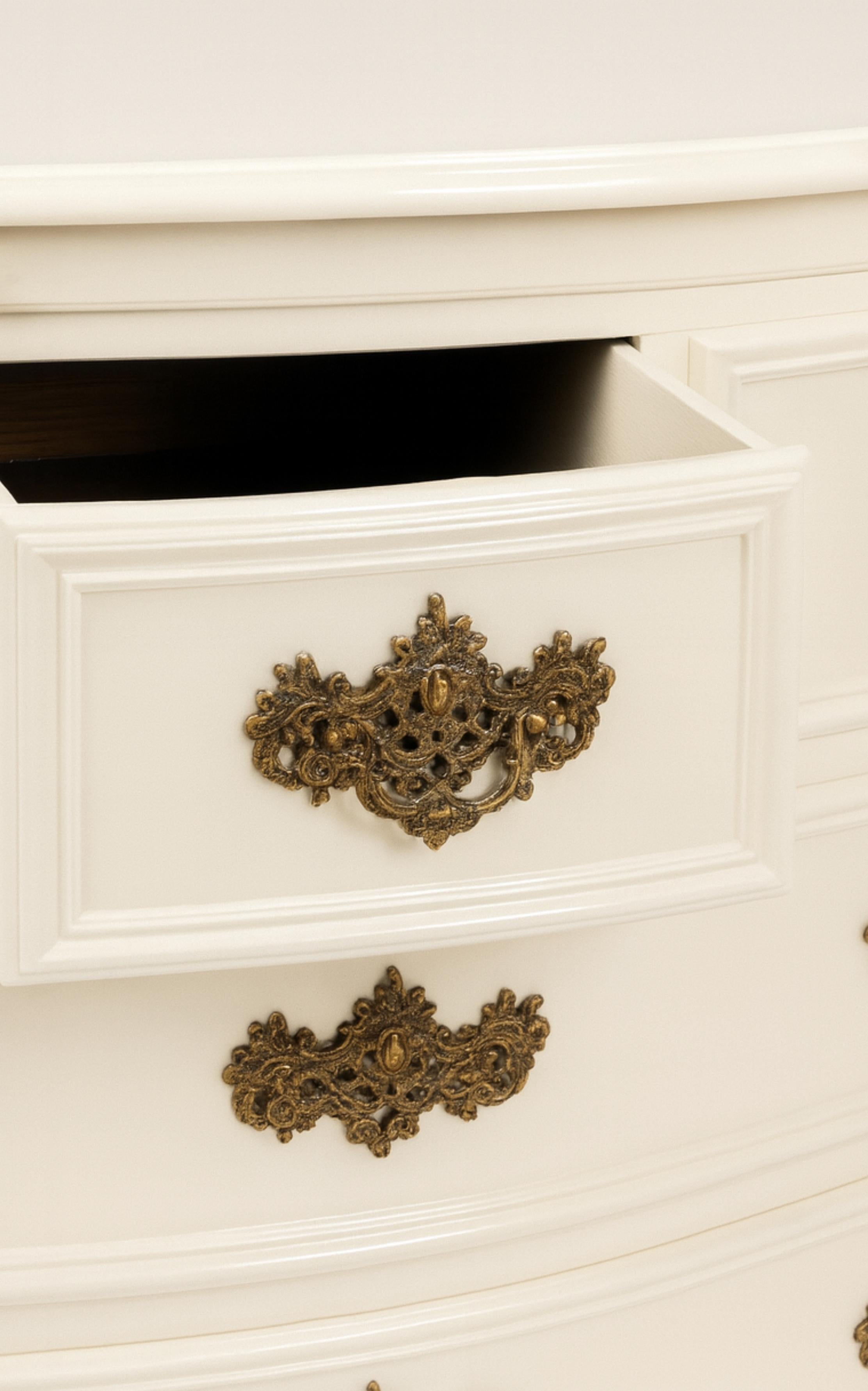 A  cómoda de madera de nogal barnizada en blanco con tapa y esquinas festoneadas, dos cajones pequeños y tres grandes, faldones recortados y finamente tallados con motivos florales y volutas con terminaciones sinuosas. Pilastras parcialmente