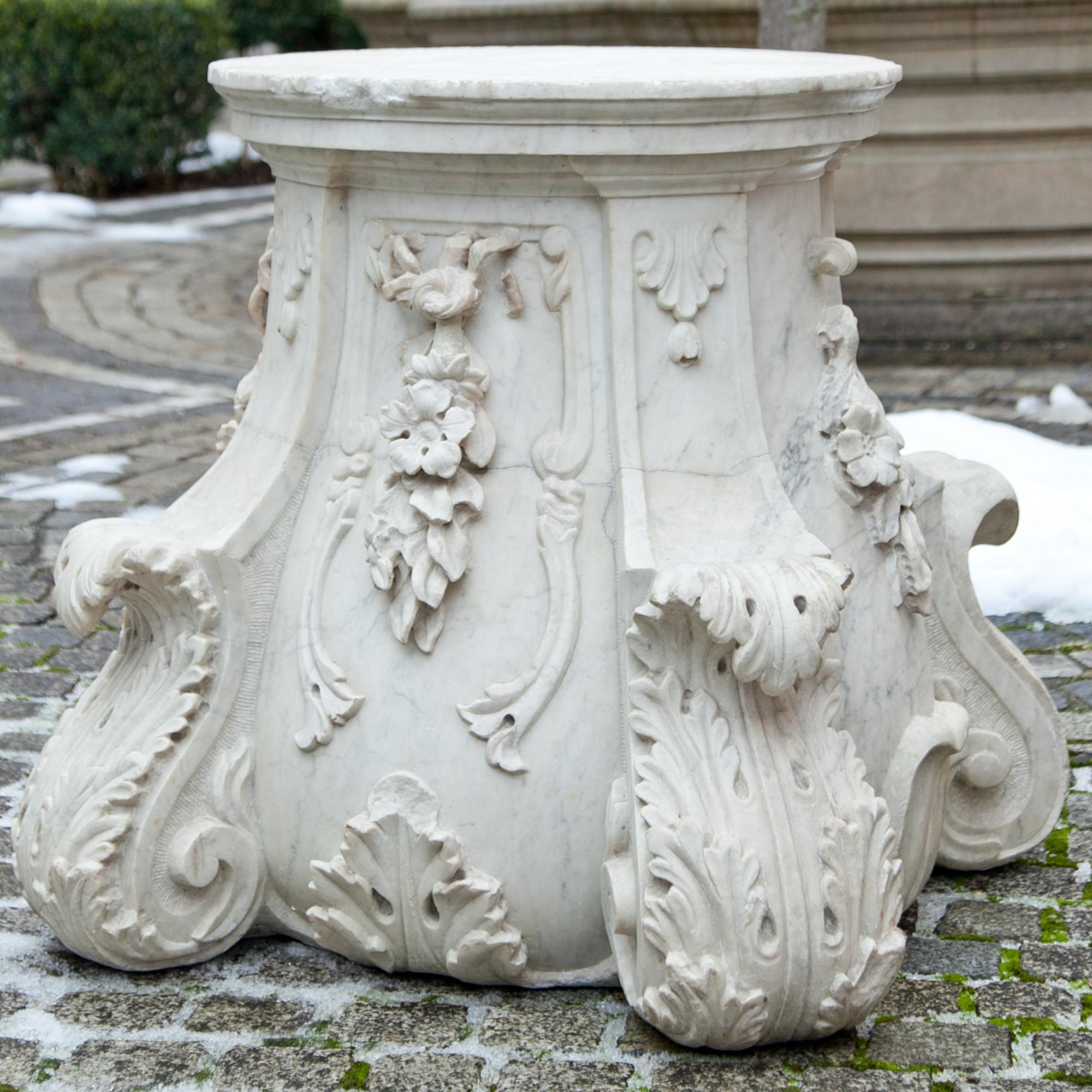 Baroque pedestal out of marble with volute corners, acanthus leaves and flower decor. Each side was carved slightly different. The round surface area is 53.5 cm in diameter and has a hole in the middle.