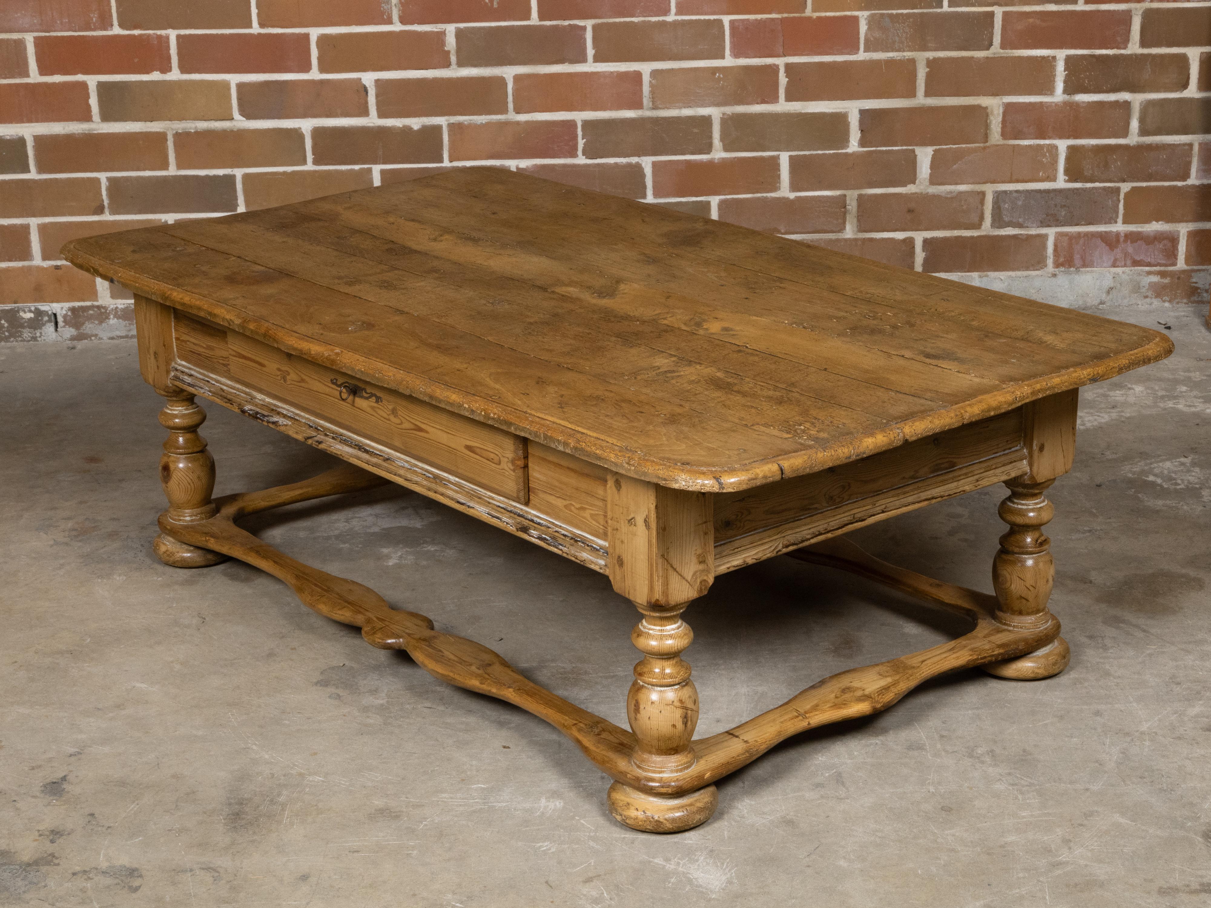 A Baroque English pine coffee table from the 19th century with turned legs, single drawer and carved side stretchers. This 19th-century Baroque English pine coffee table exudes a distinct rustic charm, highlighted by its weathered appearance and