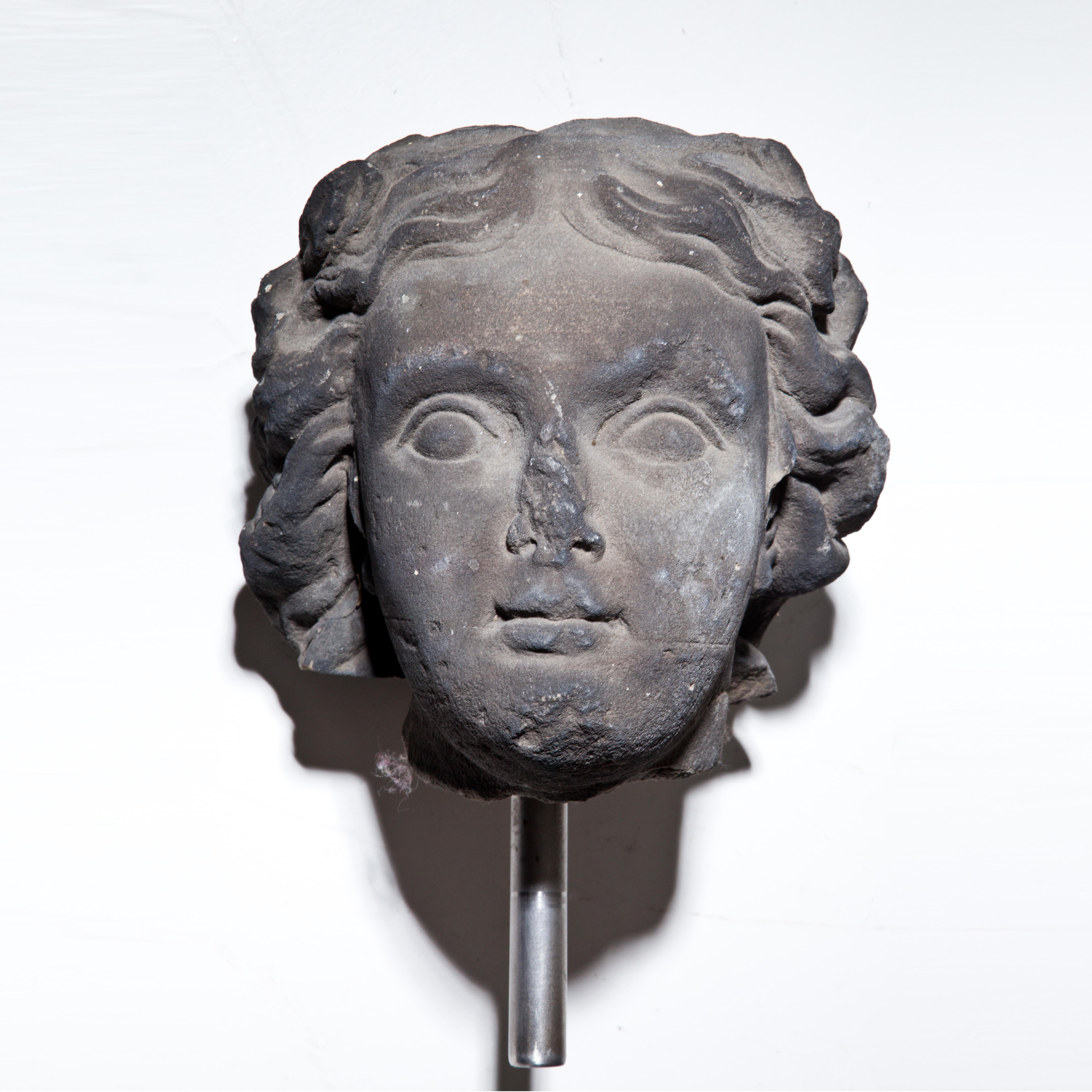 Baroque fragment of a sandstone head, hand carved with bumps on the chin, nose and the curly hair. Mounted on a metal rod. Discoloration of the stone due to weathering. Length without rod: 23 cm.