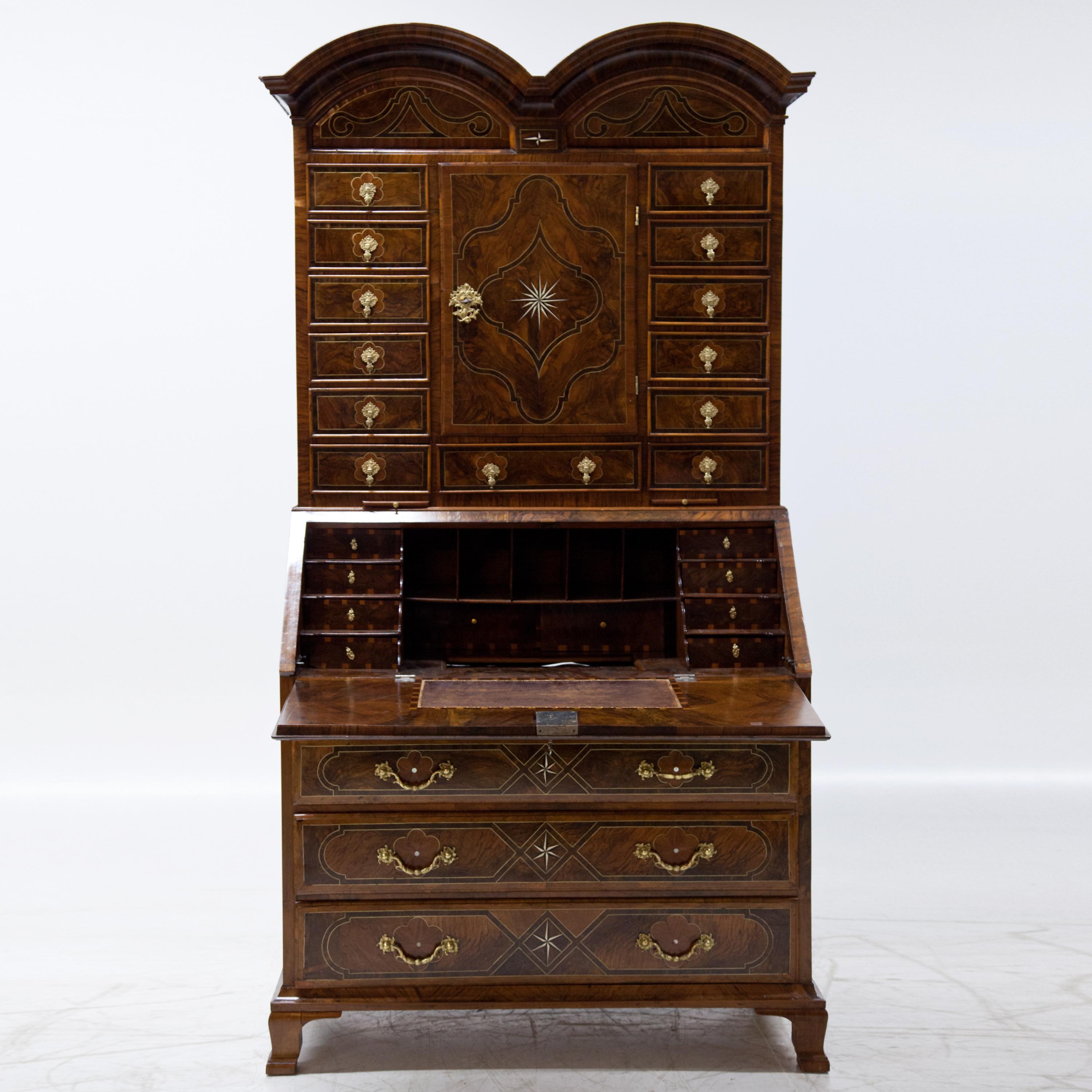 Baroque secretaire on low feet and strictly symmetrical construction. Above the chest of drawers base, there is a writing flap and two pull-out shelves. The interior division is equipped with pigeon holes and small drawers as well as a concealed