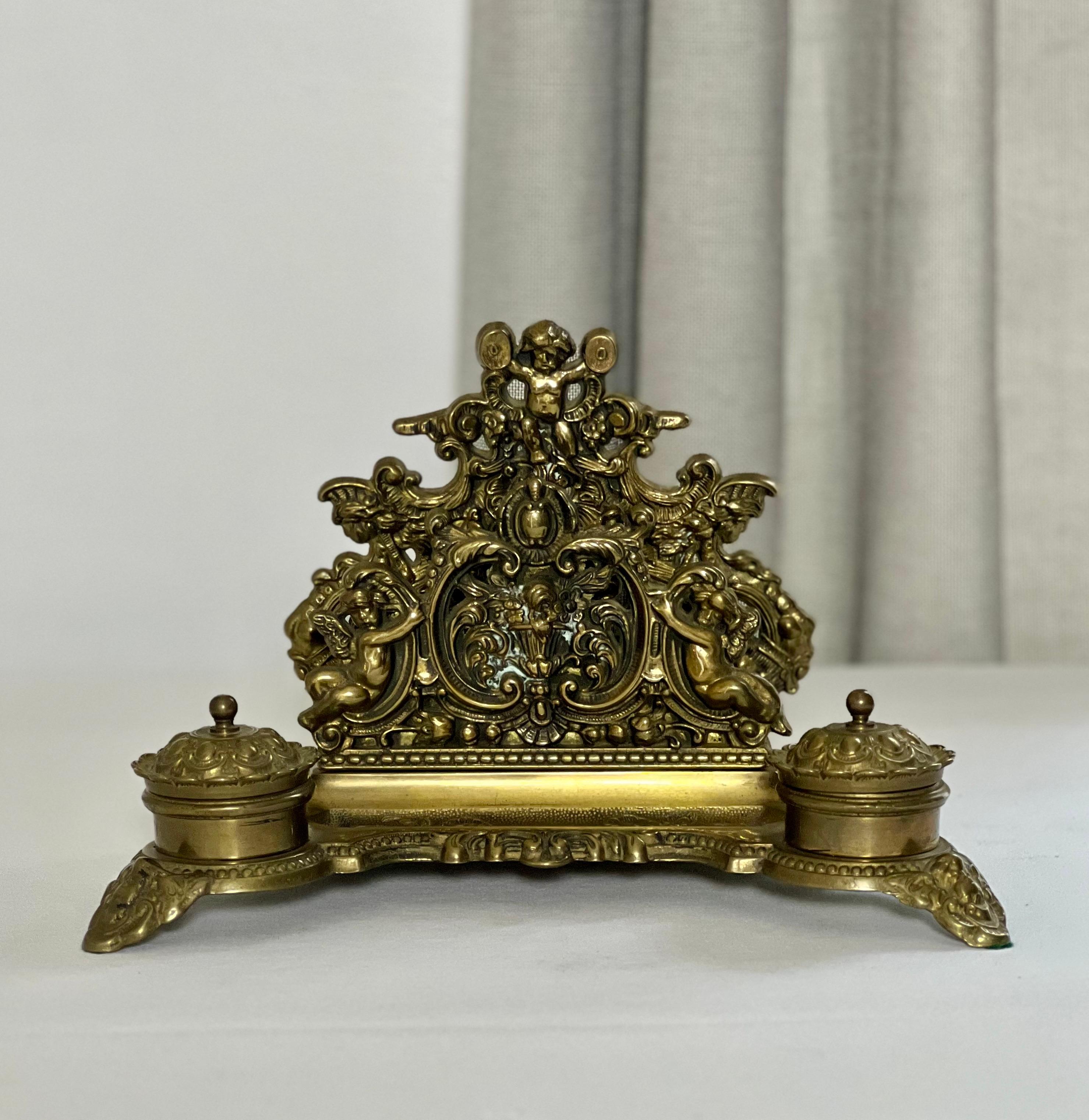 Baroque style solid brass double inkwell set with letter holder, c. 1900-1910.

A gorgeous desktop accessory ornately adorned with cherubs, flowers and scrolling acanthus leaves.  Inkwells with hinged lids and original ceramic inserts sit on either