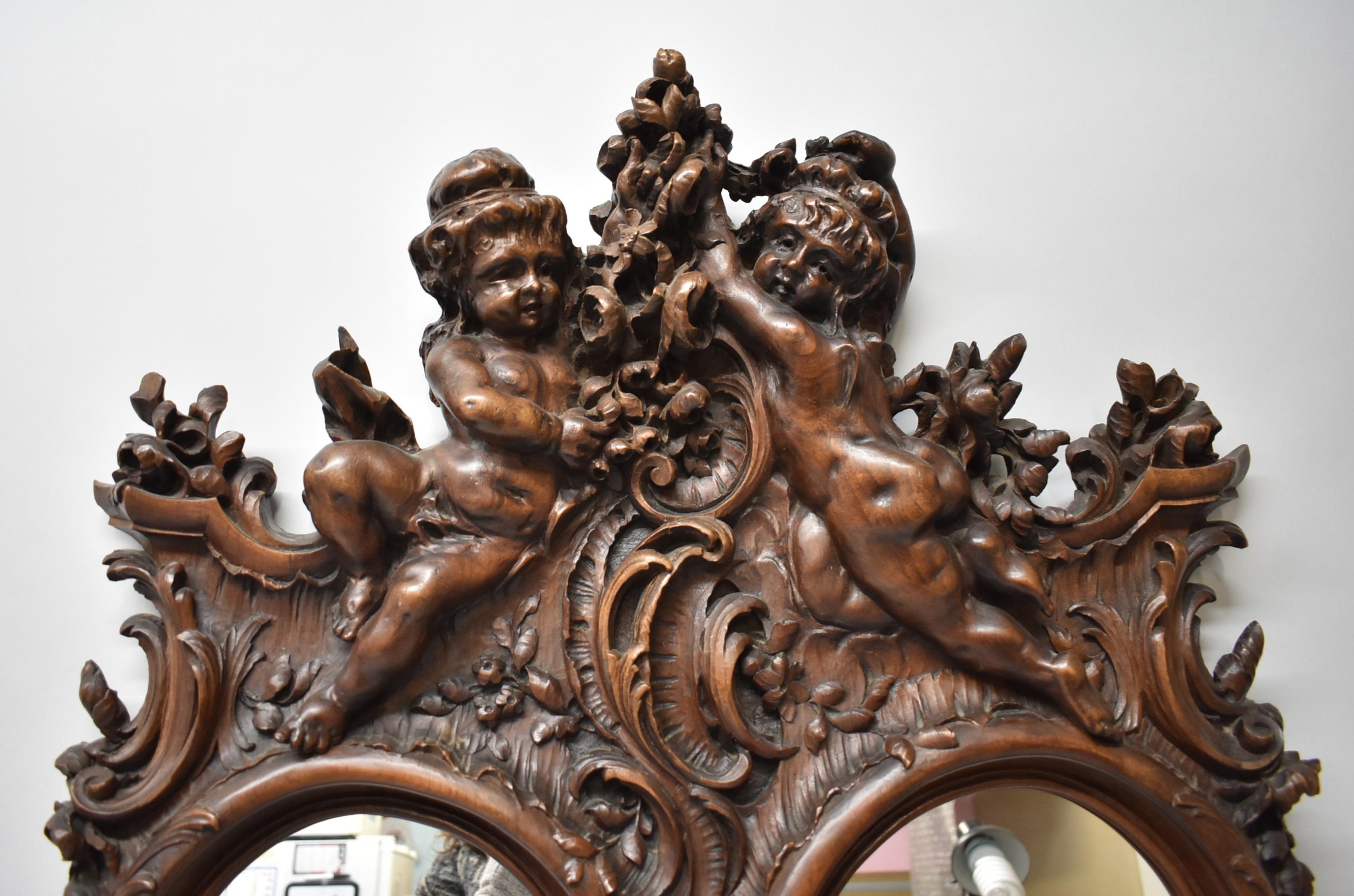 Heavily detailed carved walnut framed double oval mirror in Baroque style. Two carved cherubs grace the top and one winged cherub at the bottom of the frame. Floral details and scrolls accent the surrounding areas. Small area of carving missing as