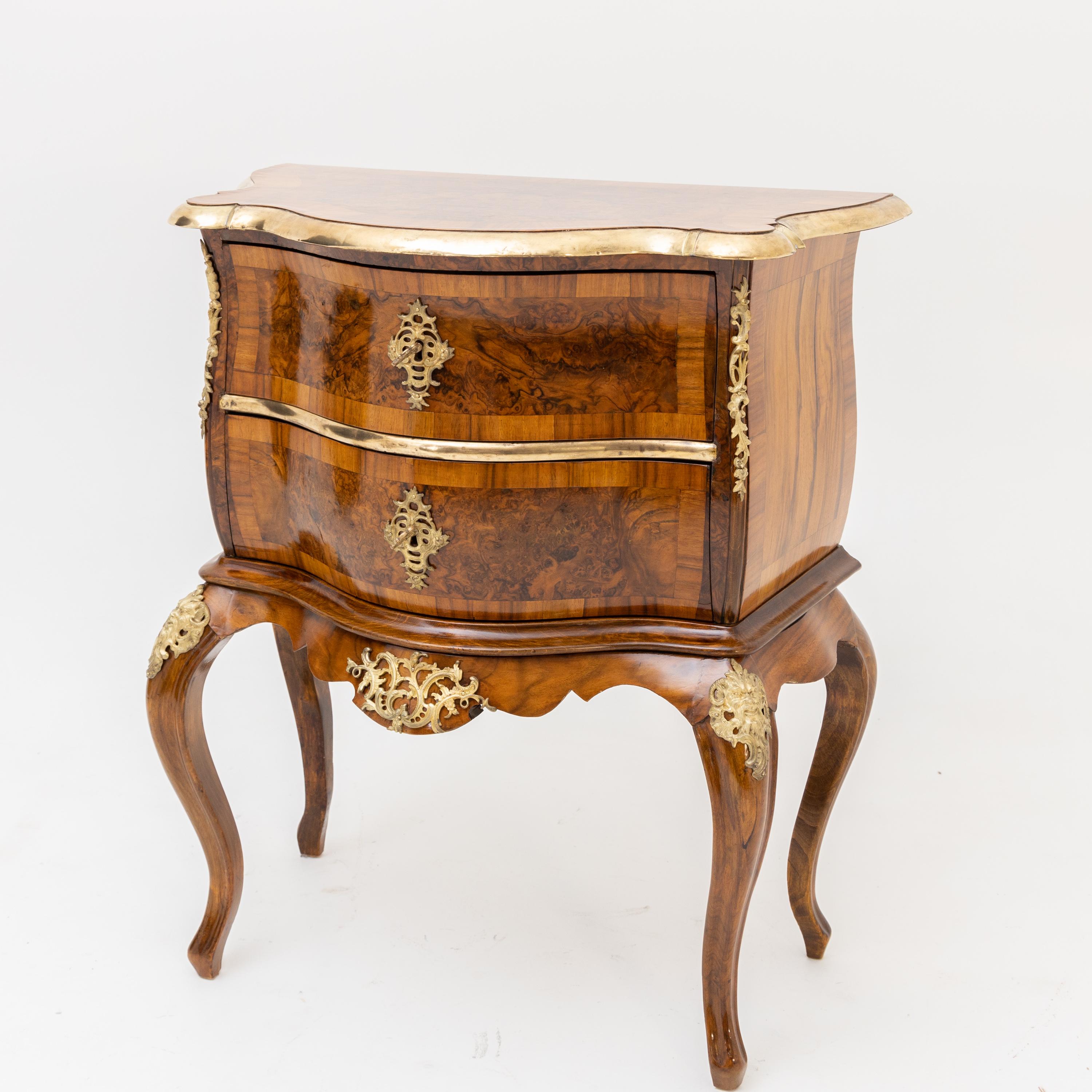 Small two-drawer chest of drawers on s-legs with pronounced shoulders and curved apron. The corpus is slightly convex and trapezoidal. The edge and crosspiece are set off in gold. Fire-gilt fittings on the front, the corners and the shoulders (here