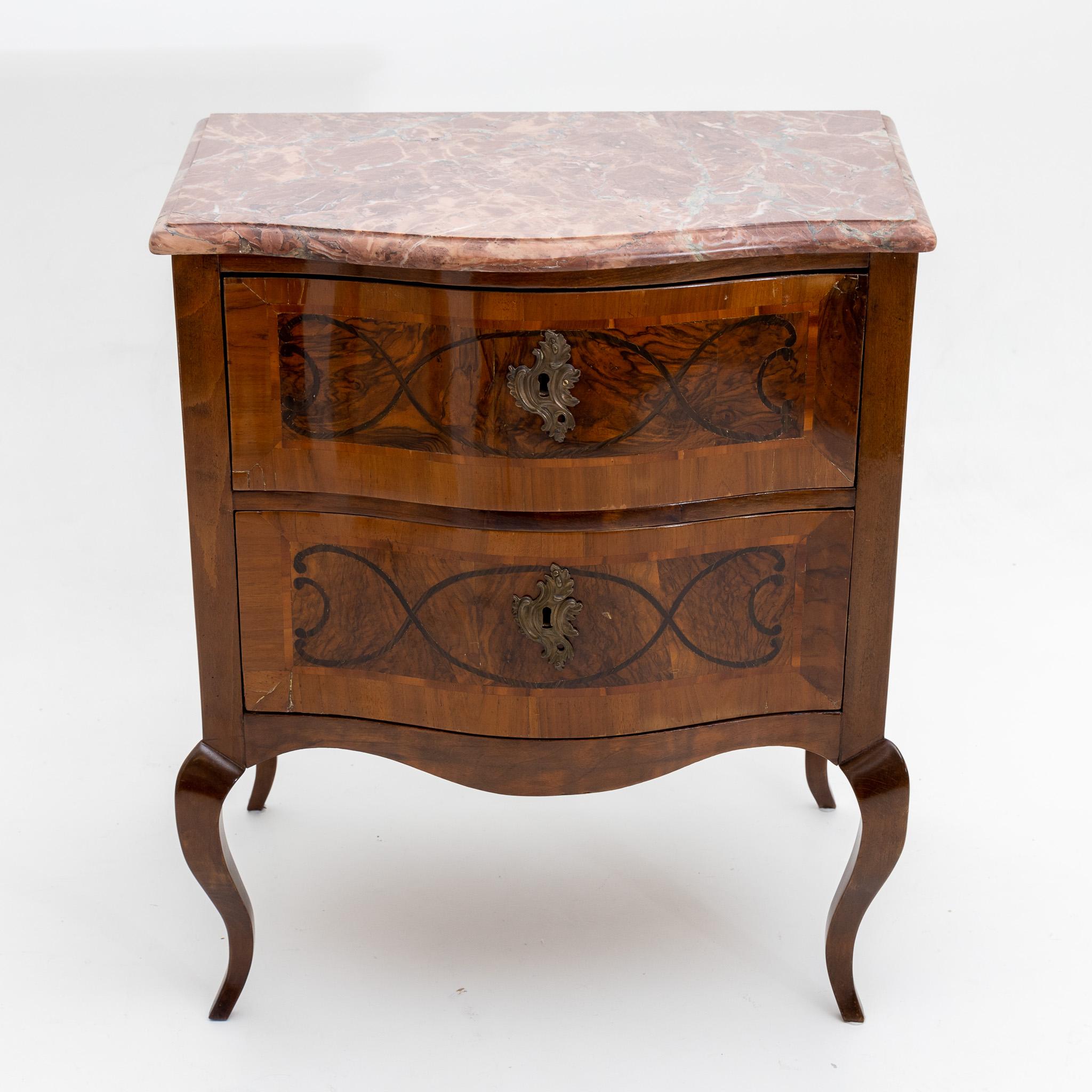 Small chest of drawers in Baroque style with two drawers, curved frame and flared S-legs. The slightly bulging front is decorated with rocaille-shaped fittings and inlaid with ribbonwork. The walnut veneered corpus is in an unrestored condition and