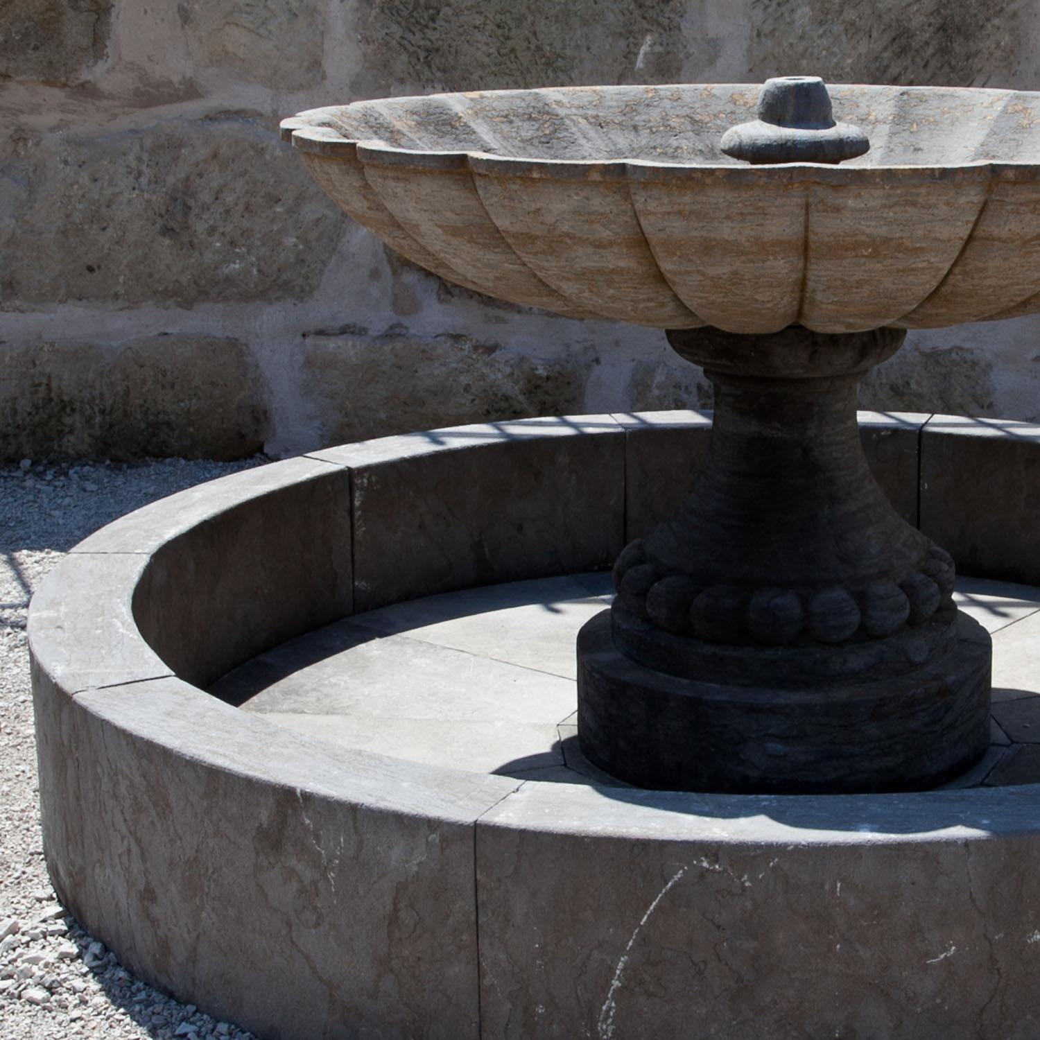 Round fountain with a short basin wall (H: 32 cm) and an extraordinary central column. The column stands on a darker baluster-shaped base and has a large scalloped basin (H: 89 x Ø140 cm). The fountain is hand-carved out of bluestone.