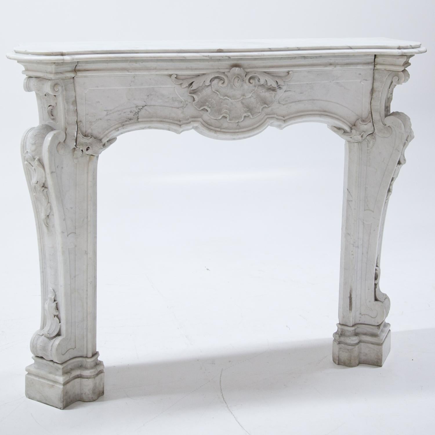 Baroque style mantel piece out of polished white marble with grey veins. The mantel is decorated with volute und leaf decor as well as a large shell ornament in the middle. The sides are at an angle and stand on profiled bases. Inner Measurements: