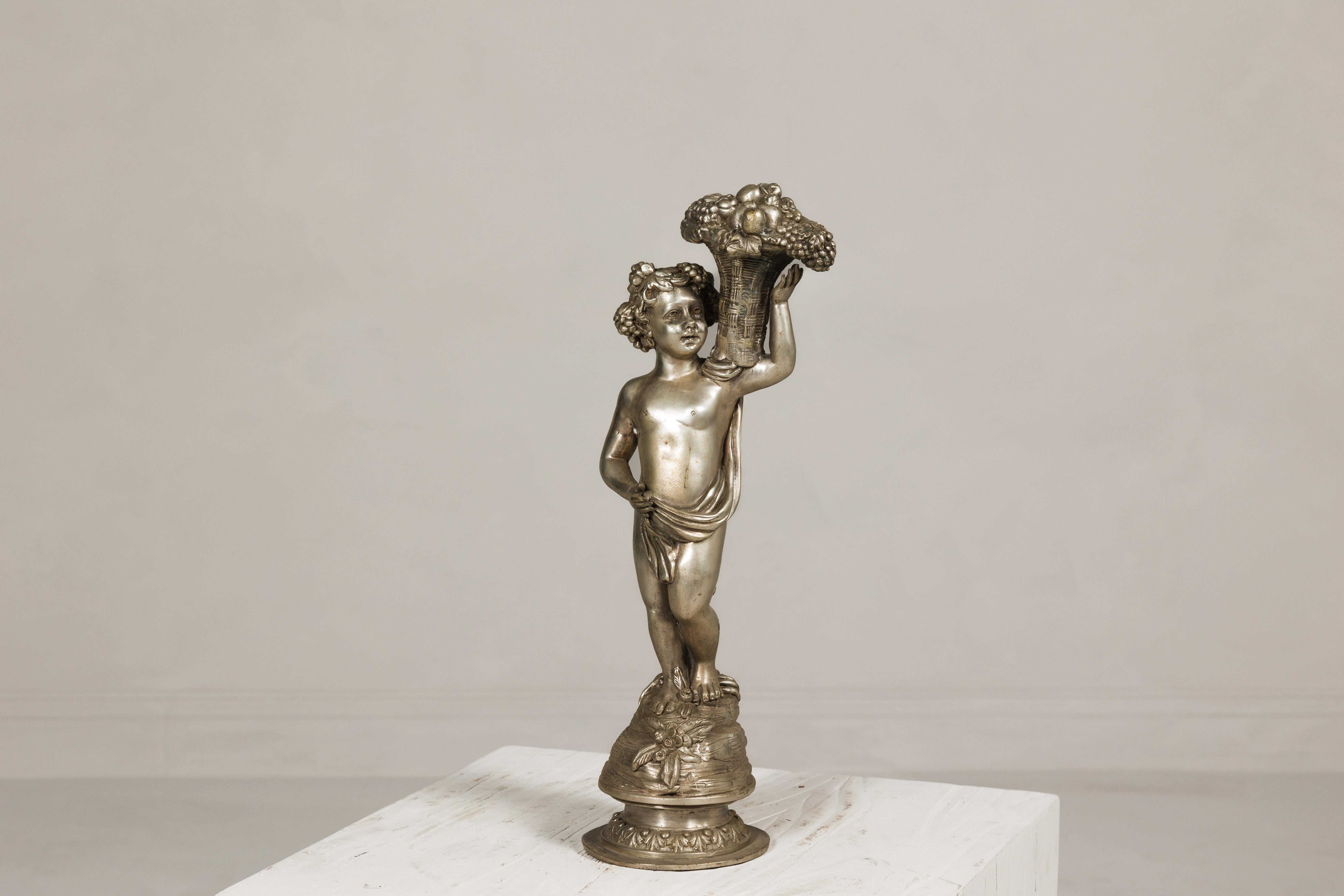 Statuetta in bronzo d'epoca in stile barocco raffigurante un putto che trasporta un cesto di frutta in vimini con patina d'argento. Ispirata al dramma e all'opulenza dell'epoca barocca, questa statuetta vintage in bronzo è uno squisito omaggio