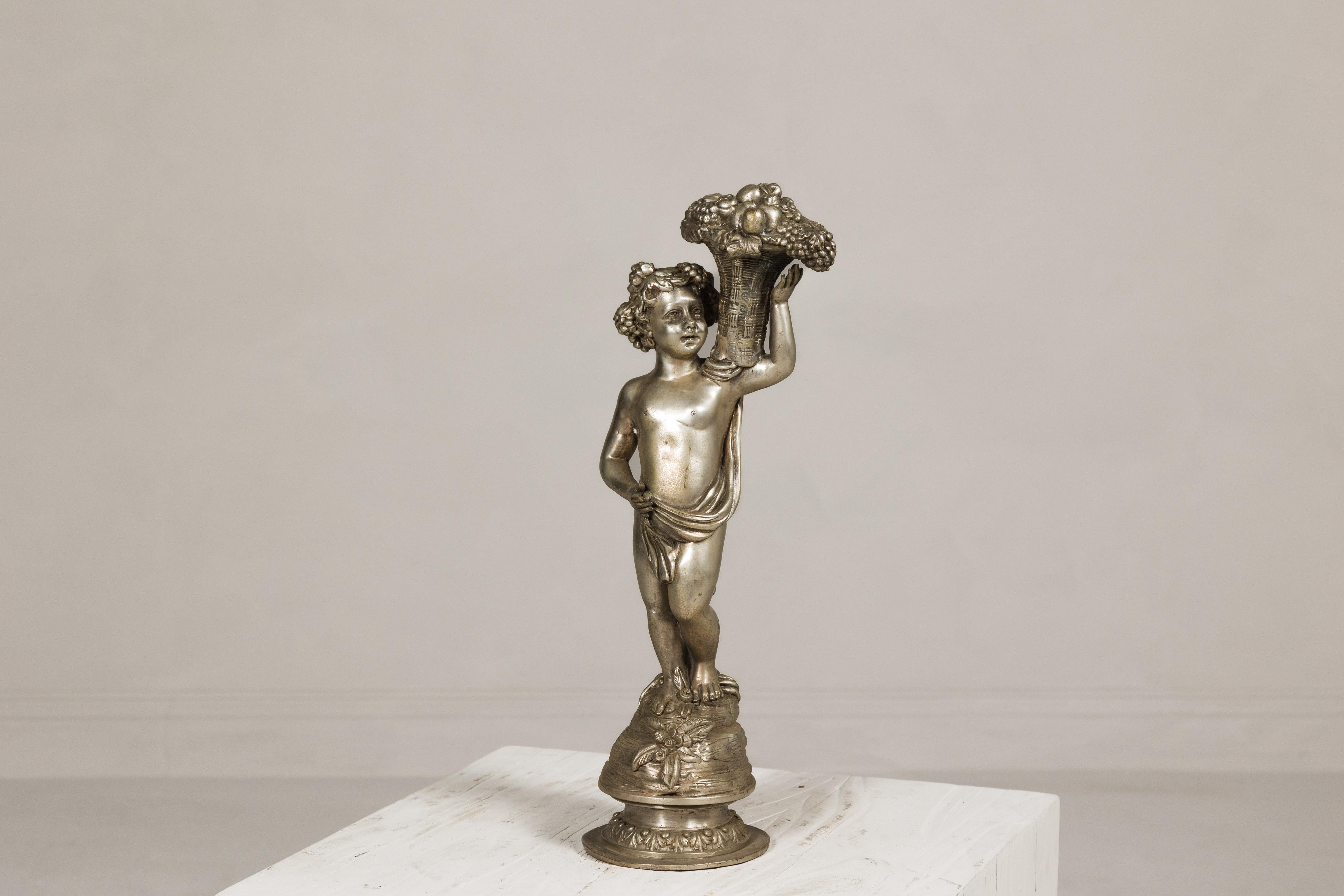 Statuette en bronze vintage de style baroque représentant un putto portant une corbeille de fruits en osier, patine argentée. Inspirée par le drame et l'opulence de l'ère baroque, cette statuette en bronze vintage est un hommage exquis à l'art