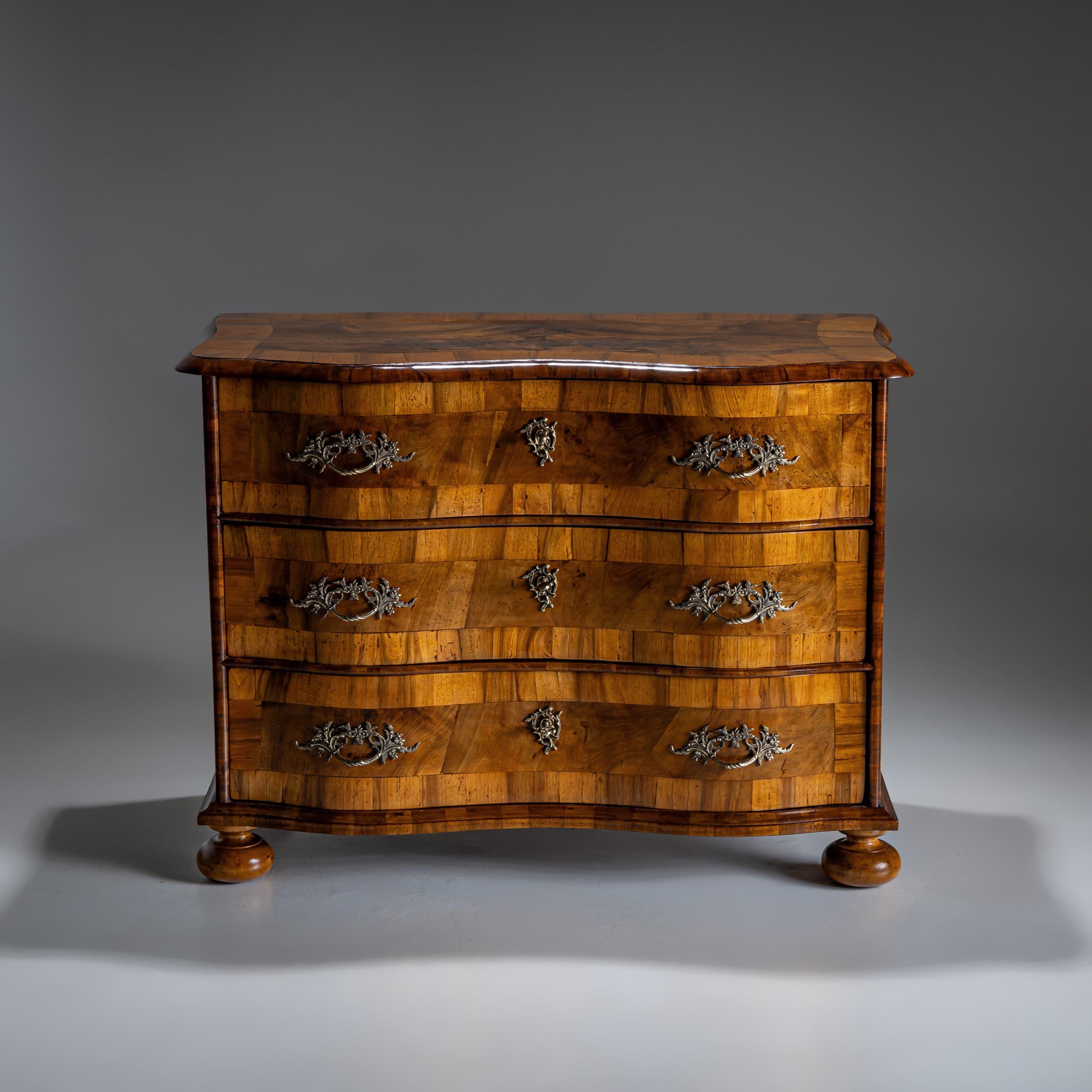Elegante commode barocco in noce con tre spaziosi cassetti e piedi a sfera compressi. Il frontale a serpentina, dolcemente curvato, è ornato da accessori asimmetrici e decorati. Traverse e pilastri arrotondati incorniciano graziosamente i cassetti,
