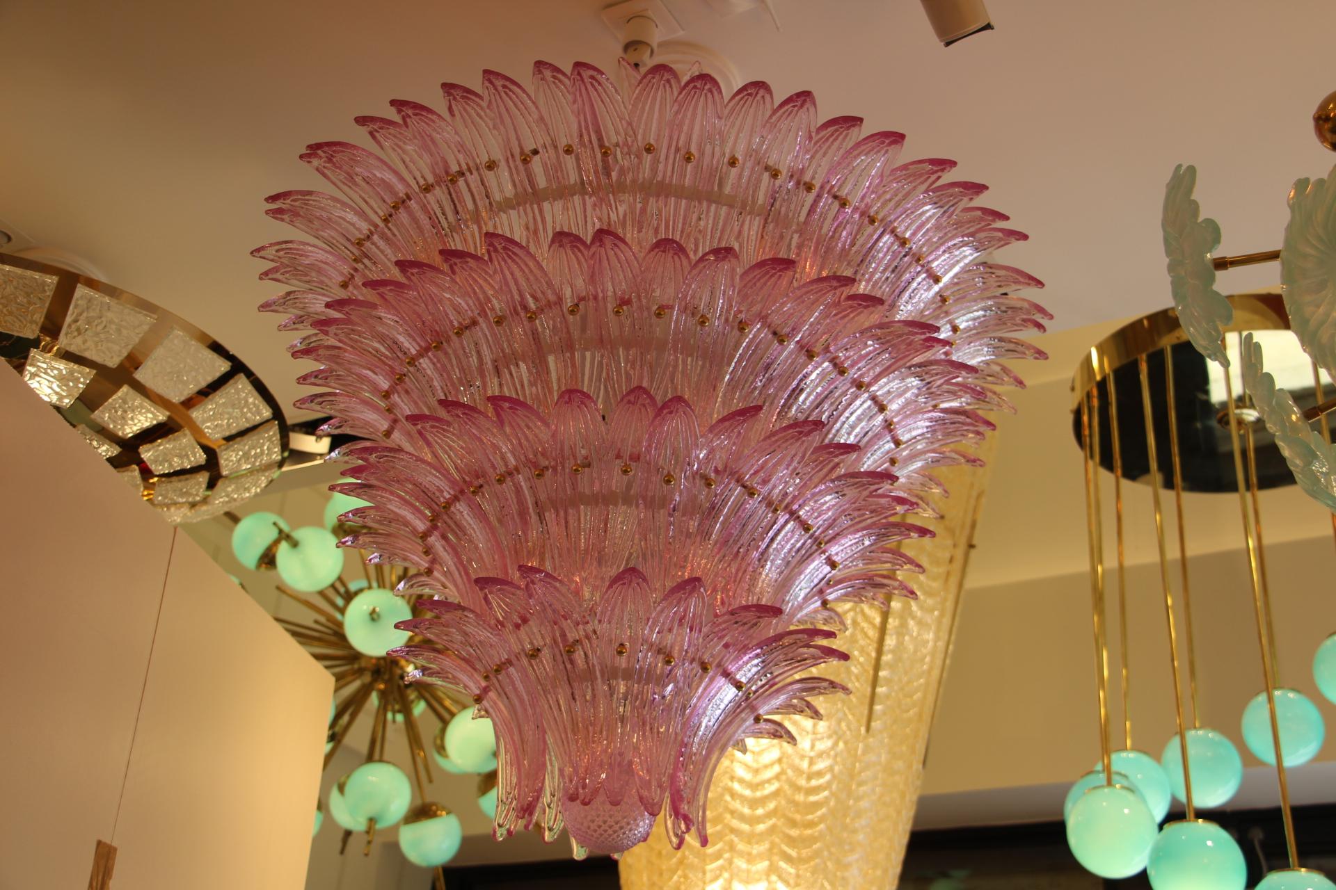 This spectacular chandelier features more than 150 hand made glass palmettes. It has got 4 tiers and a top one. Each glass palmette is fixed with a brass stud. Glass palmettes are all textured and show different nuances of pink.
Its color is