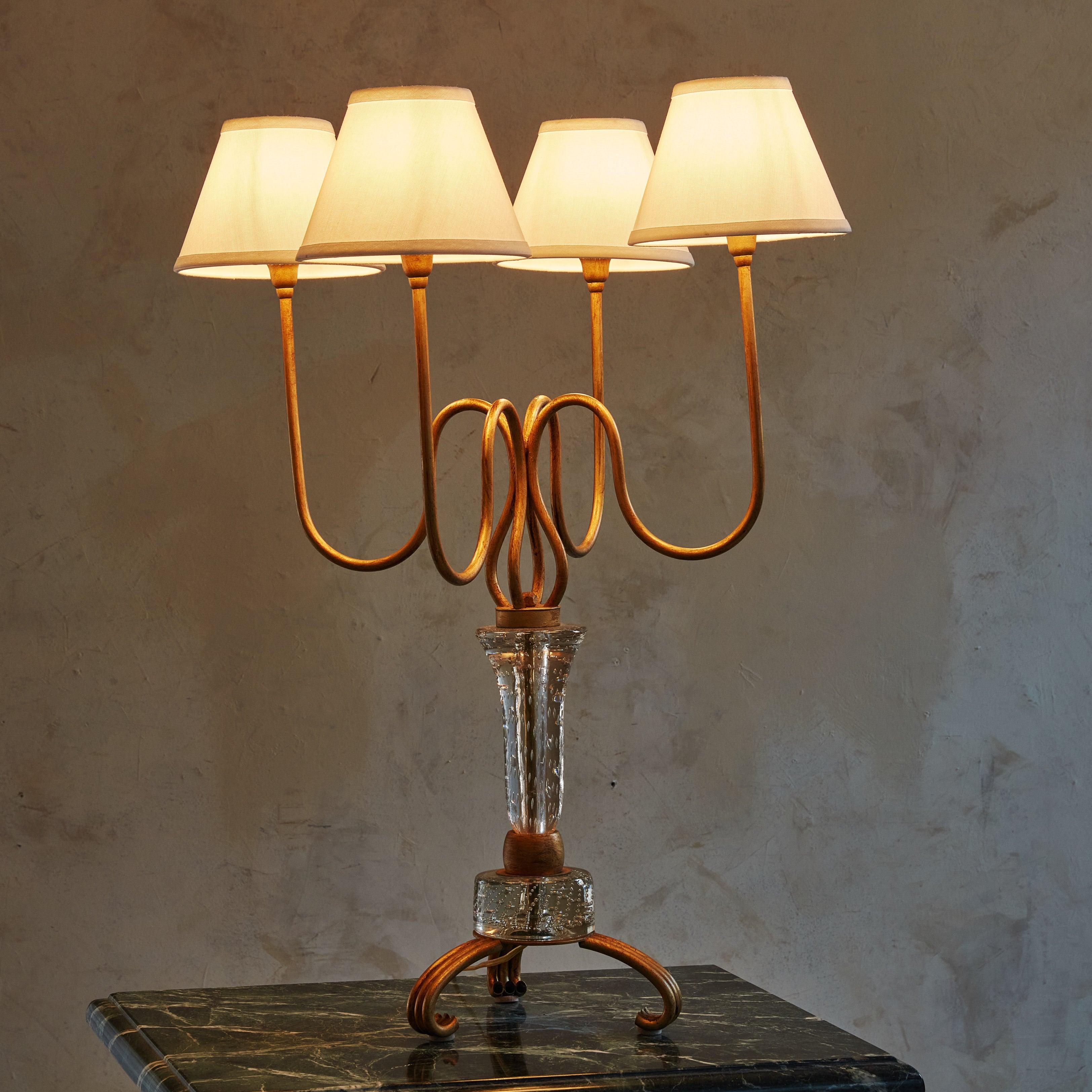 Cette lampe de table des années 1950 présente des lignes gracieuses. Dotée d'un cadre sinueux en métal doré, ses courbes fluides accueillent délicatement quatre ampoules ombragées qui diffusent une lumière chaude et ambiante. La base est ancrée par