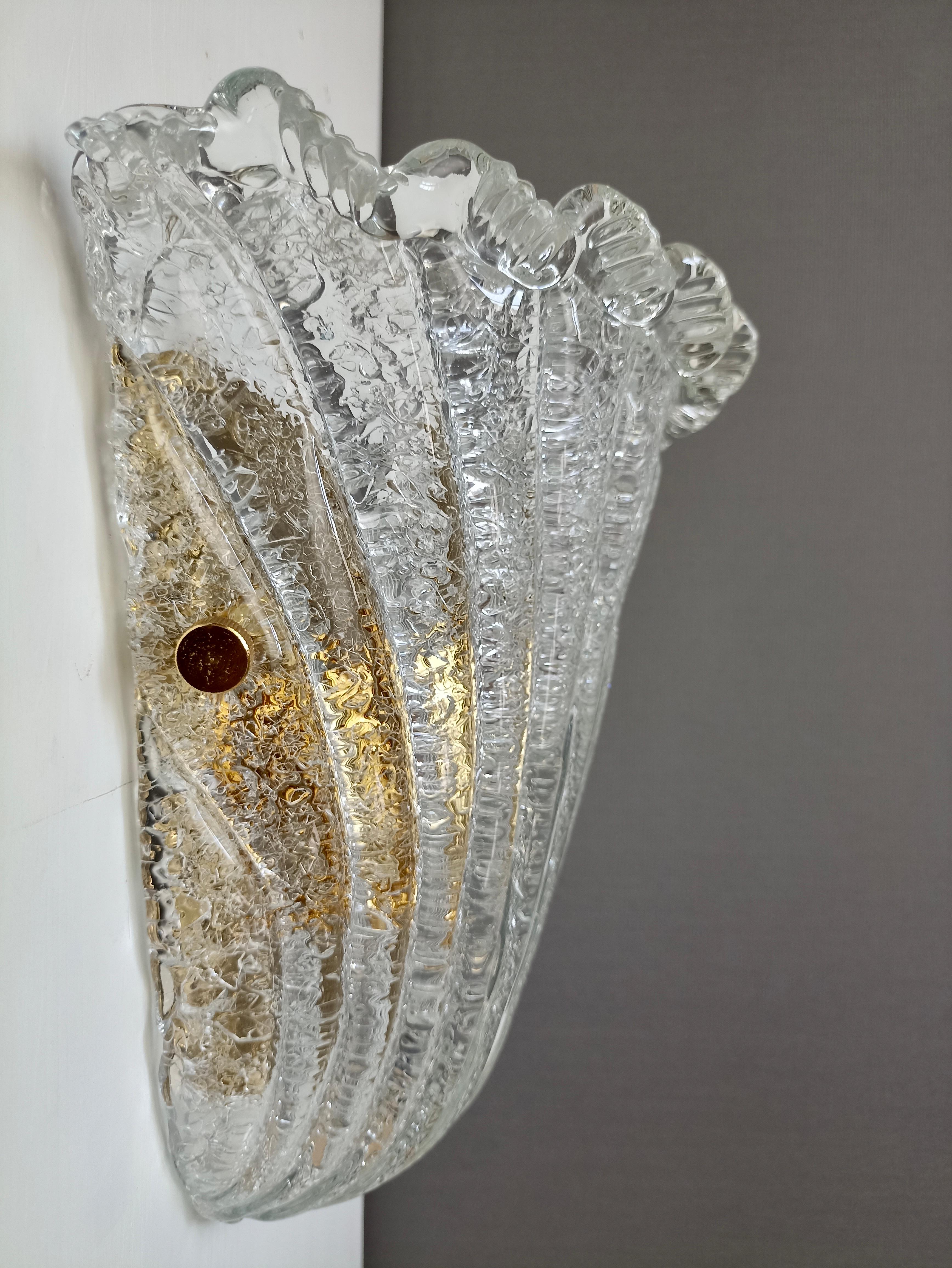 Affascinante lampada da parete a conchiglia in vetro artistico di Murano degli anni '90, nel classico stile veneziano di Barovier. 
Il paralume in vetro chiaro e liscio a coste è caratterizzato da un effetto sorprendente, ottenuto grazie