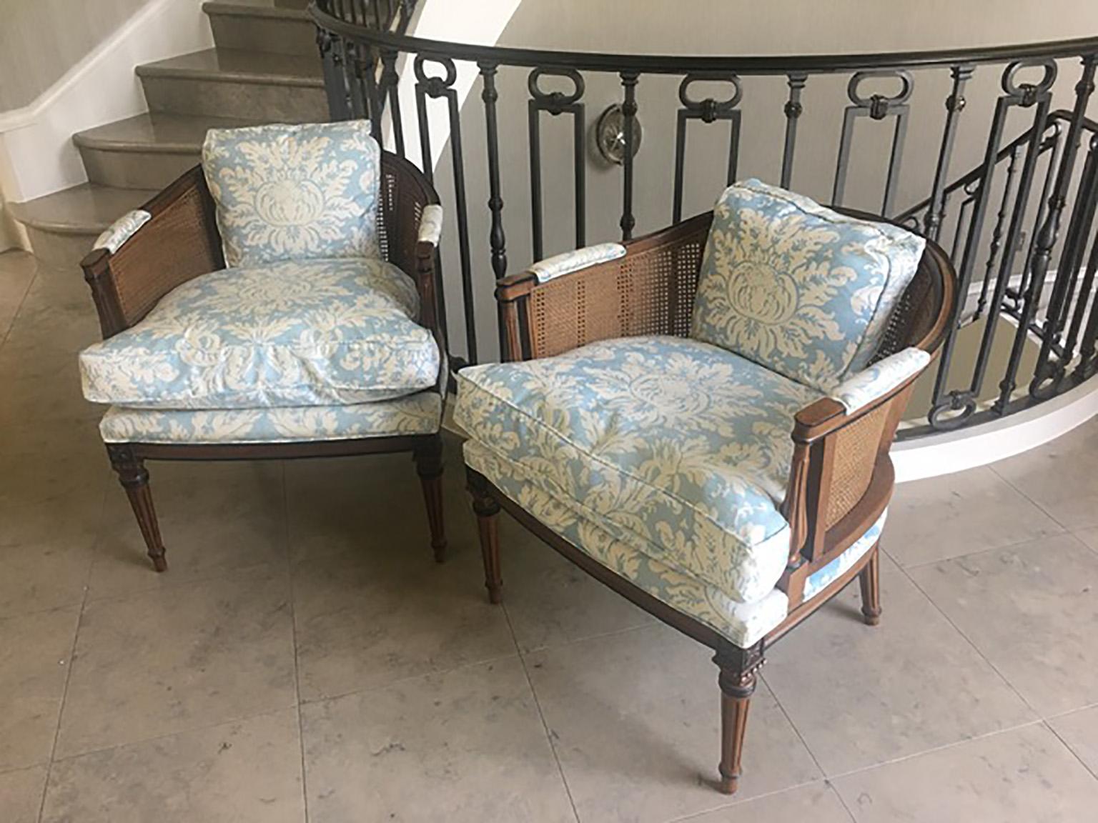 This pair of stunning vintage Hollywood Regency barrel back cane chairs is upholstered in blue silk fabric with down cushions. It is in its original finish with ebony black accents on the walnut wood frame. The cane is in great shape. The back