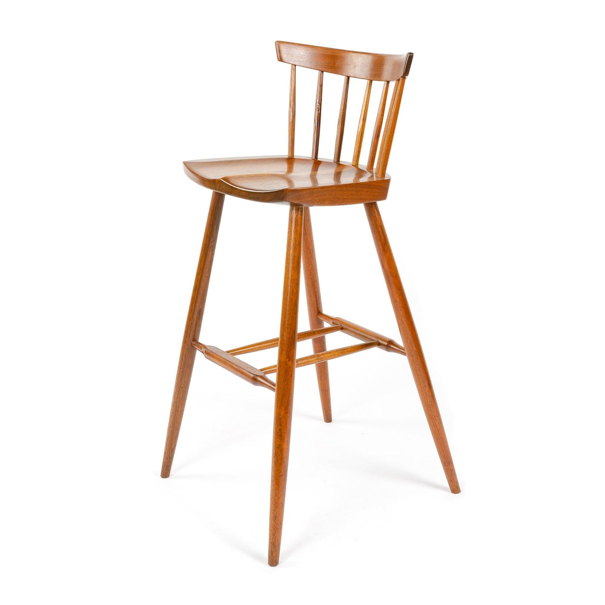 Ein Barhocker aus Nussbaumholz mit handbehauenen Spindeln, die eine dünne Rückenlehne tragen. Mit einem geformten Sitz auf vier gespreizten, sich verjüngenden Dübelbeinen mit einer Fußstützenstreckung.