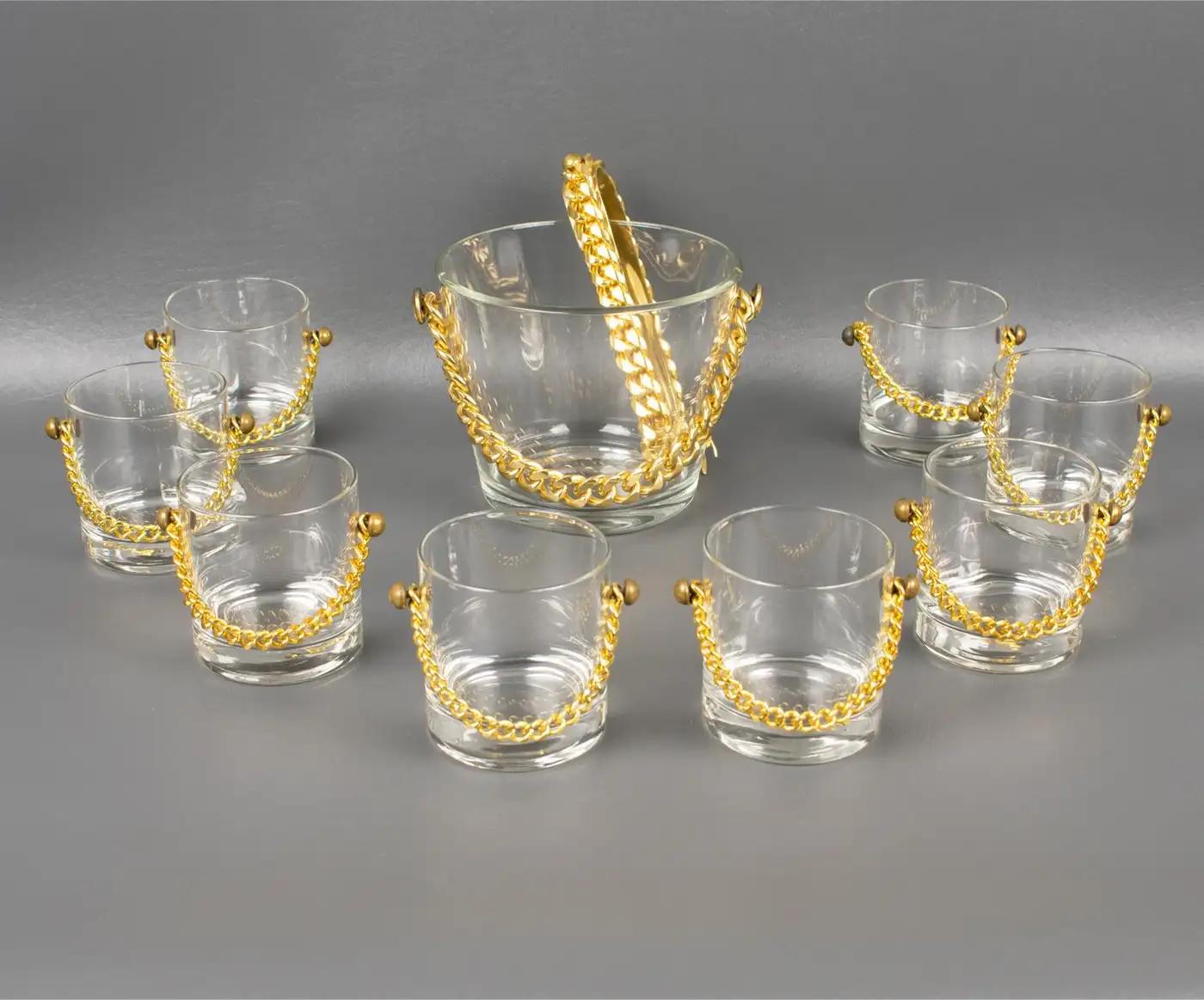 This stunning modern barware cocktail set features an ice bucket with eight matching whiskey glasses and dates from the 1980s. The incredible chic shape boasts a gilded metal chain decor on each piece. The ice bucket has a glass body with a gilded