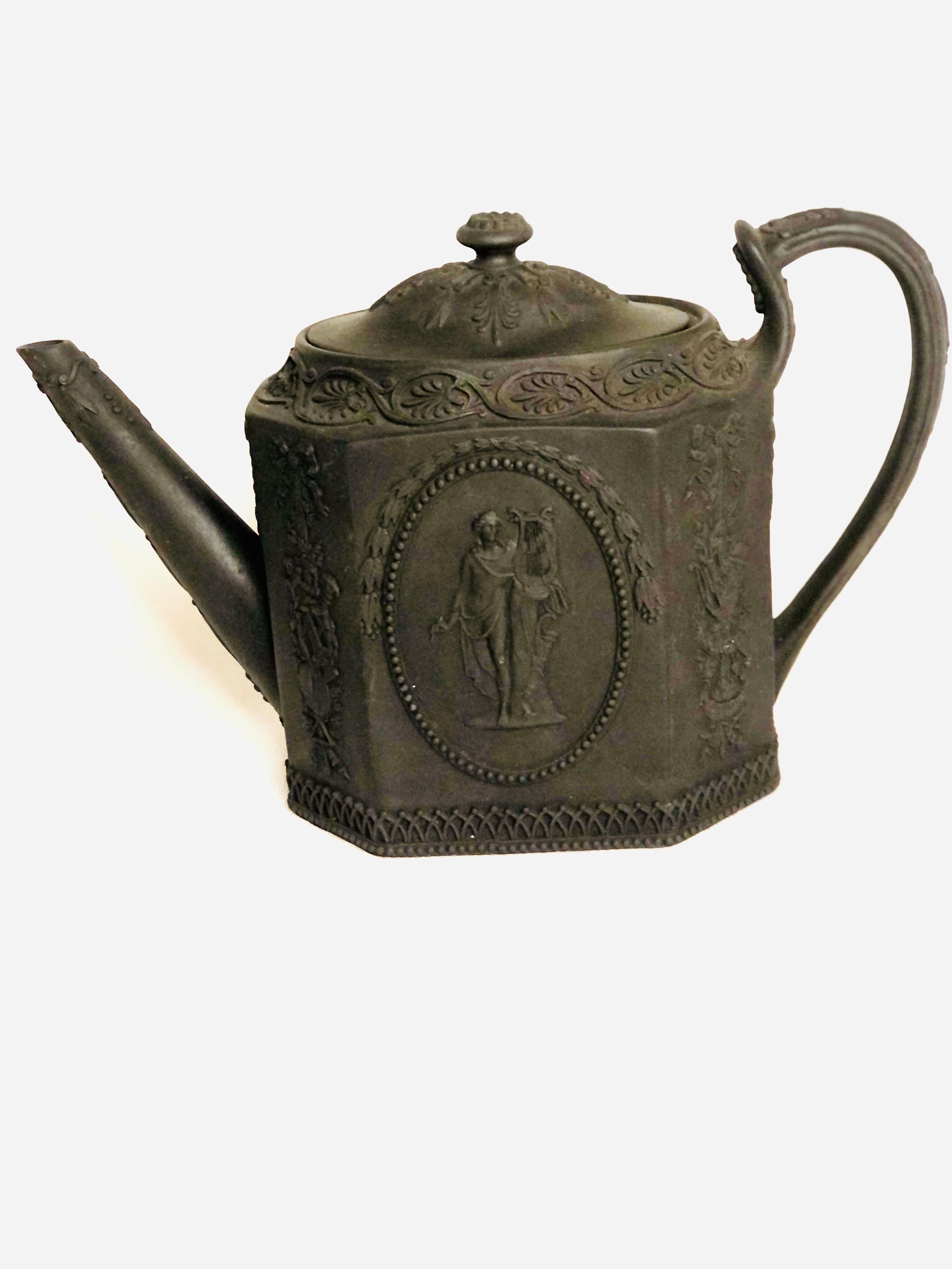 This is a very rare antique Wedgwood Etruria Basalt teapot. It is eight sided and decorated on all sides. One side has a medallion of a man playing a lyre, while the other side has a medallion of a pensive woman leaning on a plinth. There are