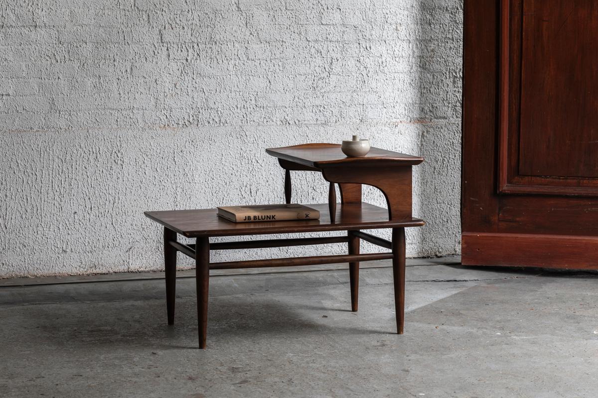 Zweistöckiger Beistelltisch, entworfen und hergestellt von Bassett Furniture in Pennsylvania in den USA um 1960. Dieser vom Art déco inspirierte Beistelltisch wurde aus Nussbaumholz gefertigt. Die nach oben geschwungenen Seiten und die sich