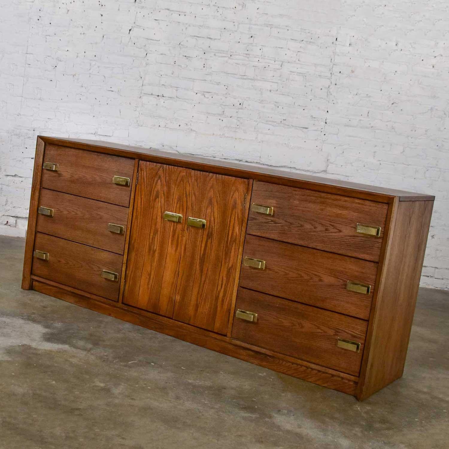 Gorgeous modern almost Brutalist Bassett credenza or buffet comprised of a medium tone finished wood (possibly oak, ash, or hickory); brass-plated die cast pulls; six drawers (three on either end), and two center doors with 3 interior drawers. In